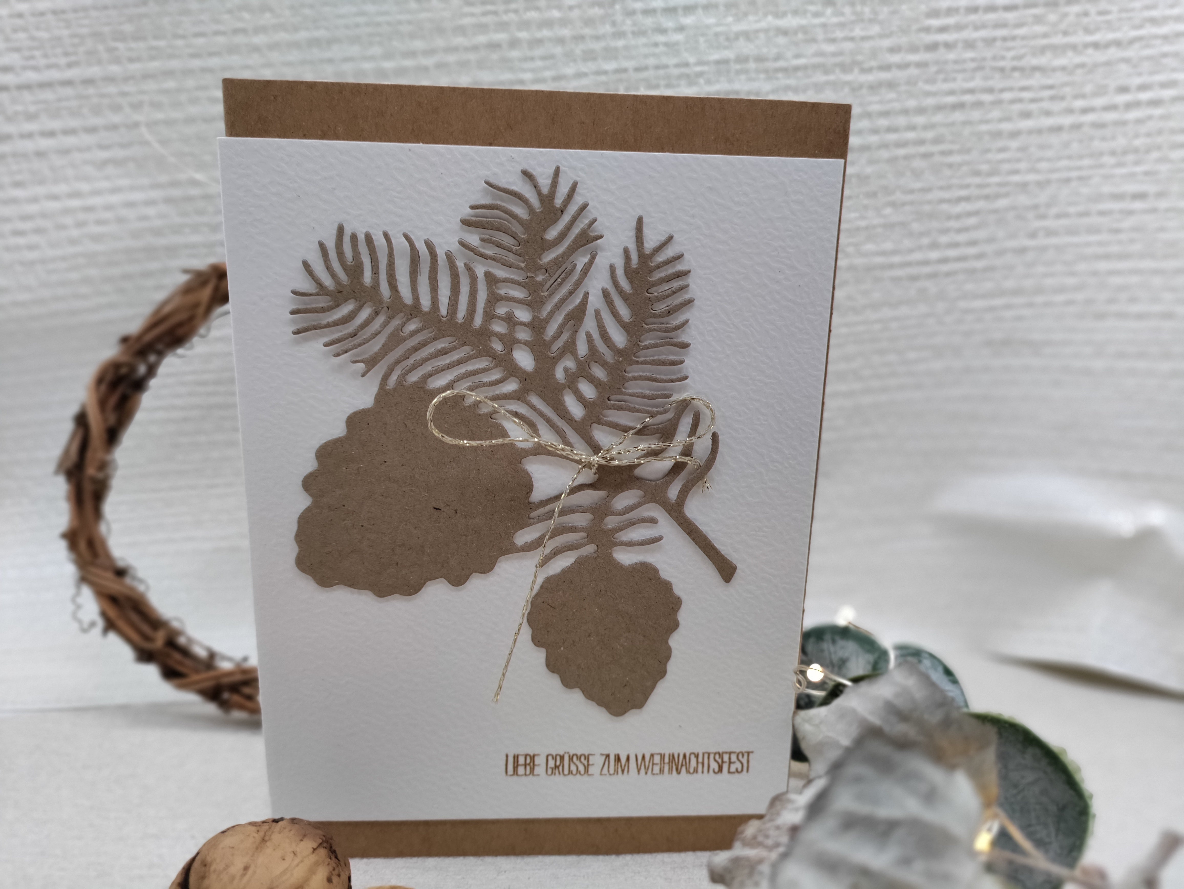 Diese handgefertigte Karte zeigt ausgeschnittene Tannenzweige und Zapfen mit der Stanzschablone: Zapfenzweig von Stanzenshop.de, verziert mit einer goldenen Schleife und dem Schriftzug "Liebe Grüße zum Weihnachtsfest" - ideal zum Basteln oder Scrapbooking.