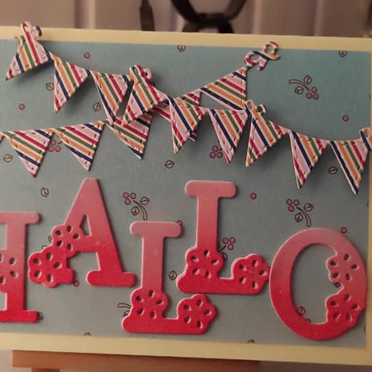 Eine handgefertigte Karte mit "HALLO" in fetten roten Buchstaben, erstellt mit der Stanzschablone: Blumenalphabet von Stanzenshop.de, zwei bunte Wimpelreihen und ein hellblauer Hintergrund, der mit kleinen Blumenmotiven verziert ist.