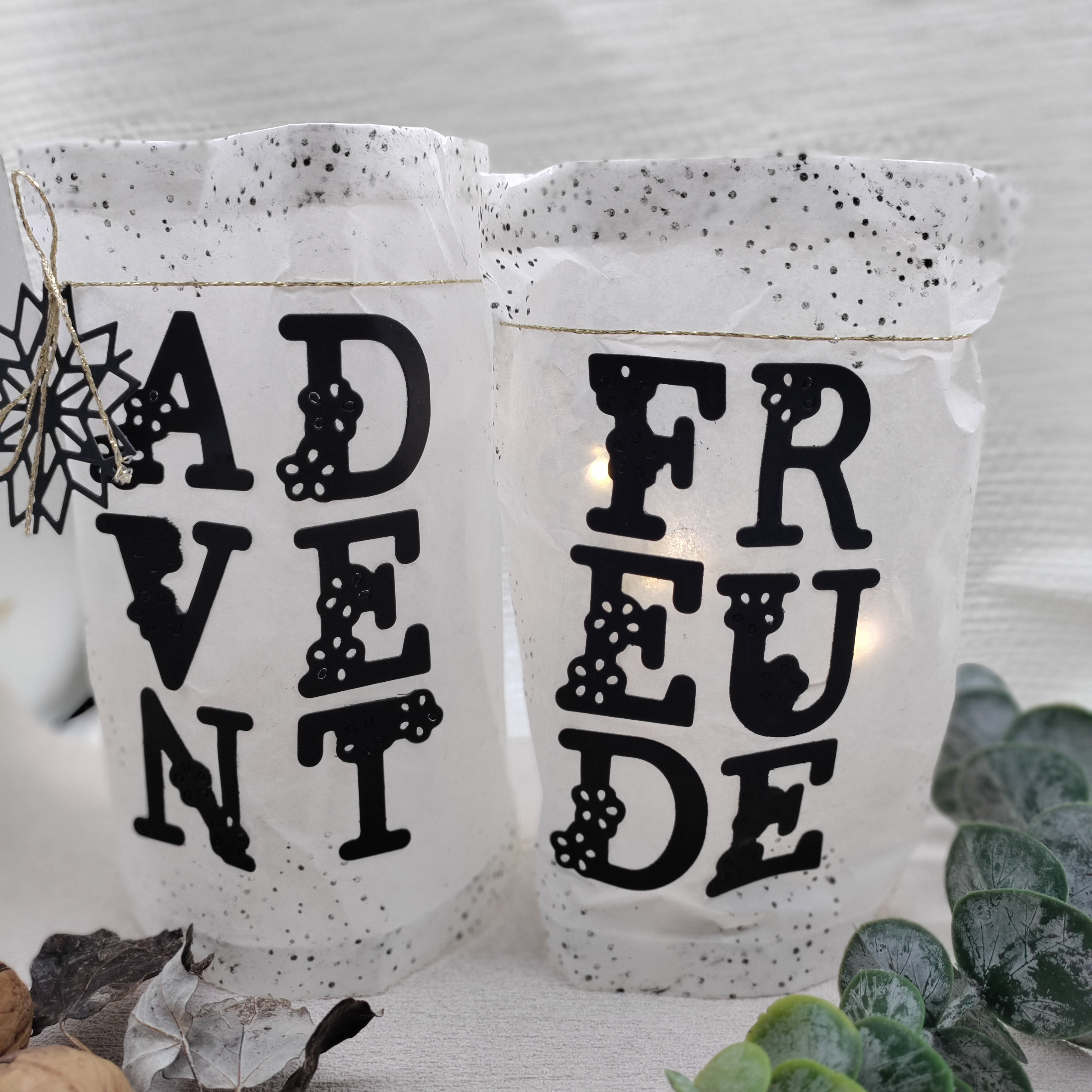 Zwei weiße Laternen mit schwarzen Buchstaben "ADVENT" und "FREUDE", geschnitten mit Stanzschablone: Blumenalphabet von Stanzenshop.de, umrahmt von Walnüssen, Blättern und Grünzeug - ideal für personalisierte Botschaften in Ihrer Einrichtung.