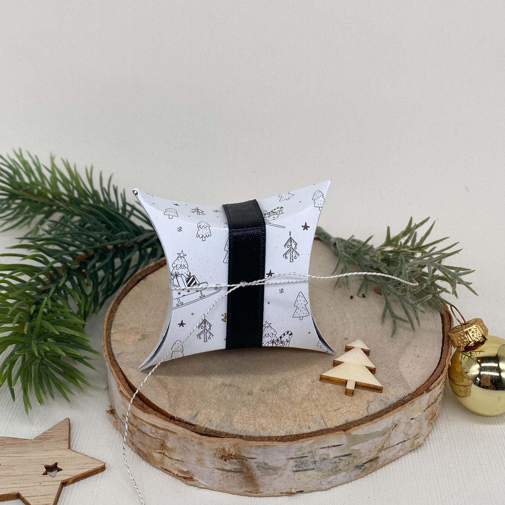 Eine Stanzschablone: Kissenschachtel von Stanzenshop.de mit schwarz-weißen Weihnachtsmotiven und einer schwarzen Schleife ruht auf Holz, umgeben von Tannenzweigen, Holzdekor und einem goldenen Ornament.