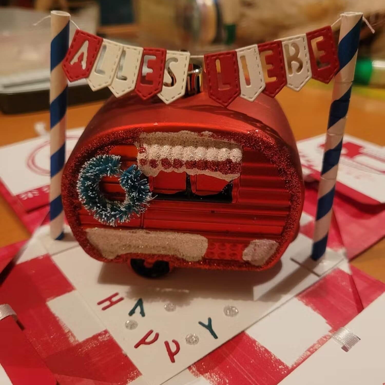 Ein rotes Ornament in Form einer Karawane mit Kranz und Schnee sitzt unter einem "ALLES LIEBE"-Banner. Für die Szene wurde die "Stanzschablone" von Stanzenshop.de verwendet: Wimpelkette mit Buchstaben", ideal für persönliche Kartendekorationen und Botschaften.