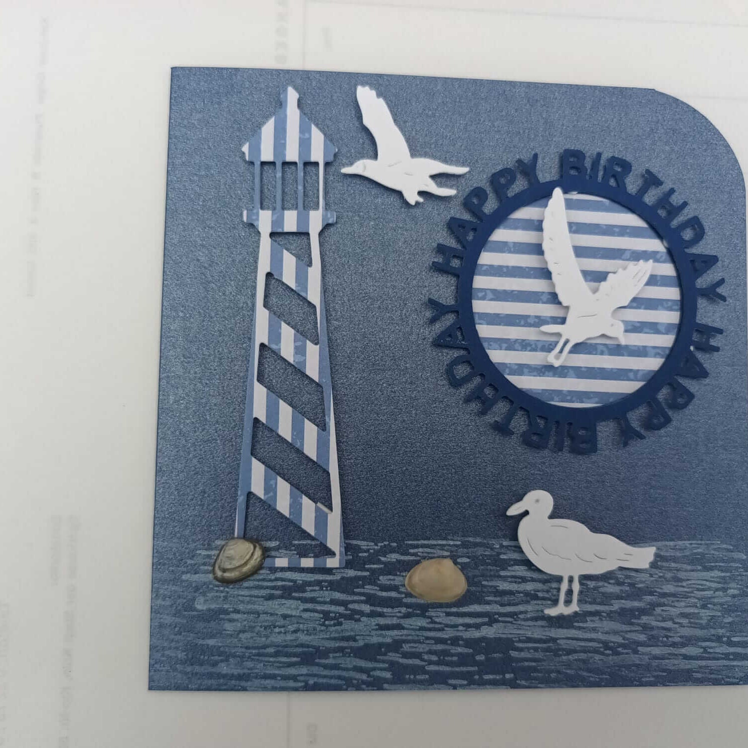 Eine blaue Geburtstagskarte mit einem gestreiften Leuchtturm, Kieselsteinen und Möwen, inspiriert von der Stanzschablone Drei Möwen von Stanzenshop.de, mit "HAPPY BIRTHDAY" in erhabenen Buchstaben um einen kreisförmigen Ausschnitt - perfekt für Scrapbooking.