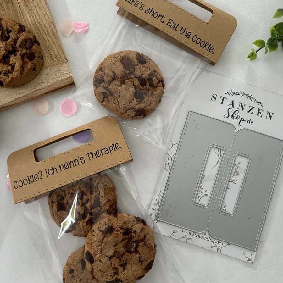 Zwei durchsichtige Tüten mit Schokoladenkeksen und Pappauflegern - auf der einen steht "Life's short. Iss den Keks." und der andere auf Deutsch - sitzen neben einer Stanzschablone Griffe von Stanzenshop.de, ideal zum Basteln oder Ausstellen Ihres Bastelergebnisses.