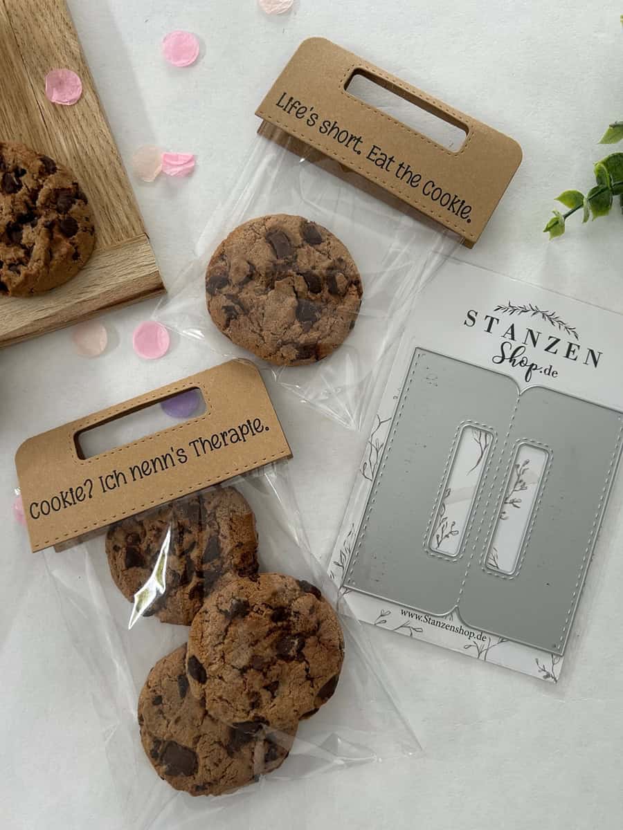 Zwei durchsichtige Tüten mit Schokoladenkeksen und Pappauflegern - auf der einen steht "Life's short. Iss den Keks." und der andere auf Deutsch - sitzen neben einer Stanzschablone Griffe von Stanzenshop.de, ideal zum Basteln oder Ausstellen Ihres Bastelergebnisses.