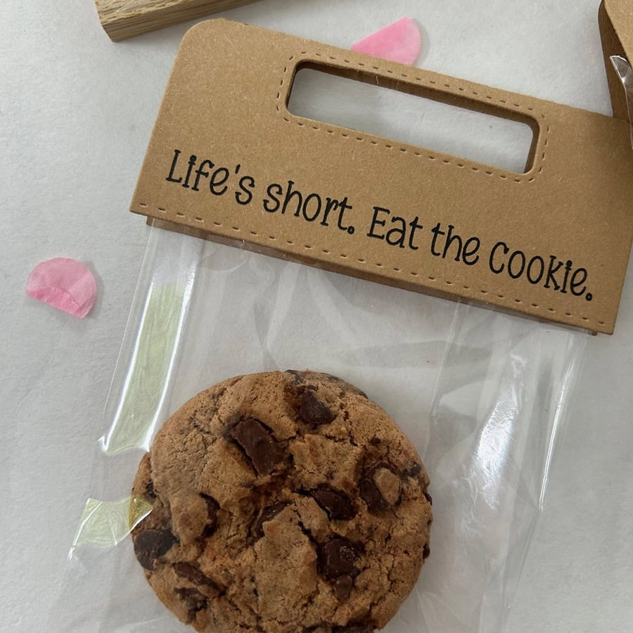 Ein Schokoladenkeks in einer durchsichtigen Tüte mit braunem Stanzschablone-Griff von Stanzenshop.de mit der Aufschrift "Life's short. Eat the Cookie". Rosa Blütenblätter und Scrapbooking-Akzente verleihen der Szene einen kreativen Touch.