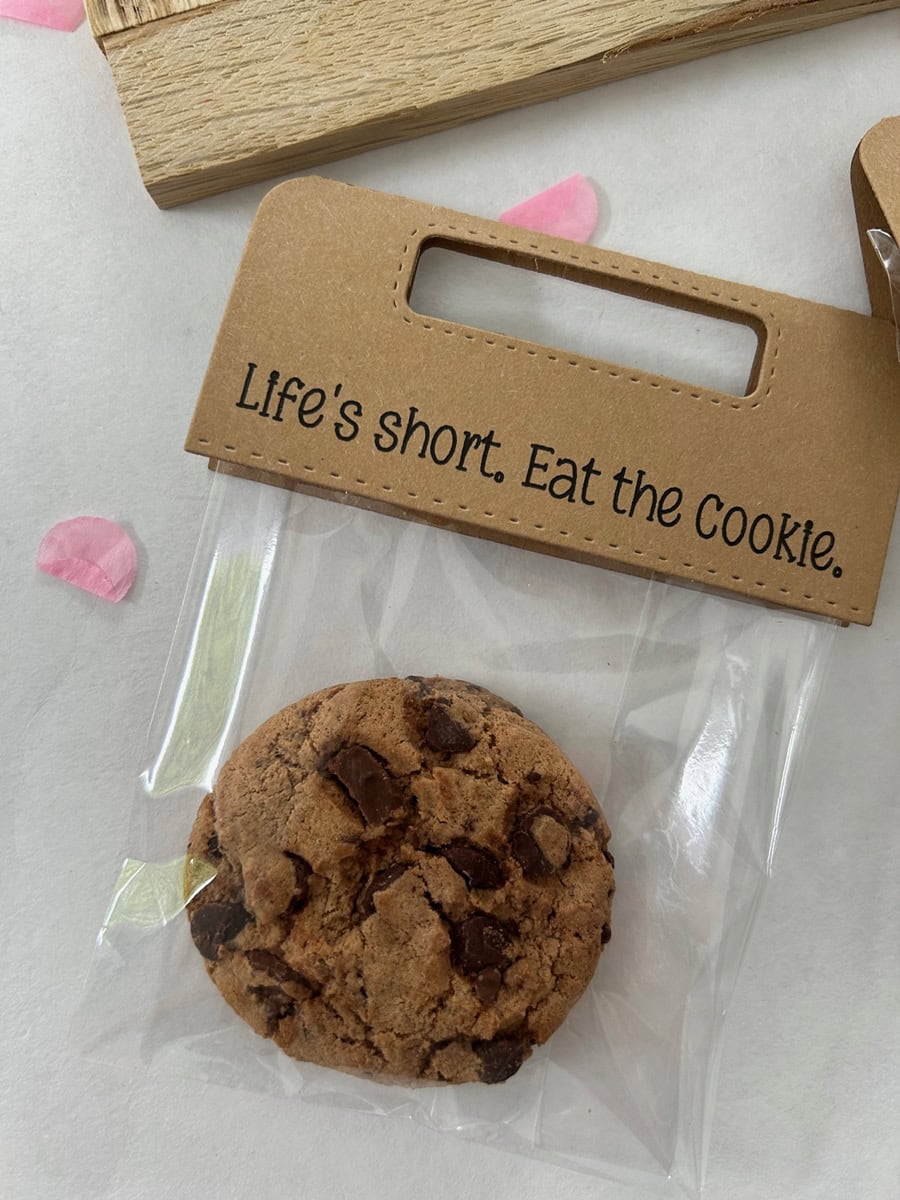 Ein Schokoladenkeks in einer durchsichtigen Tüte mit braunem Stanzschablone-Griff von Stanzenshop.de mit der Aufschrift "Life's short. Eat the Cookie". Rosa Blütenblätter und Scrapbooking-Akzente verleihen der Szene einen kreativen Touch.
