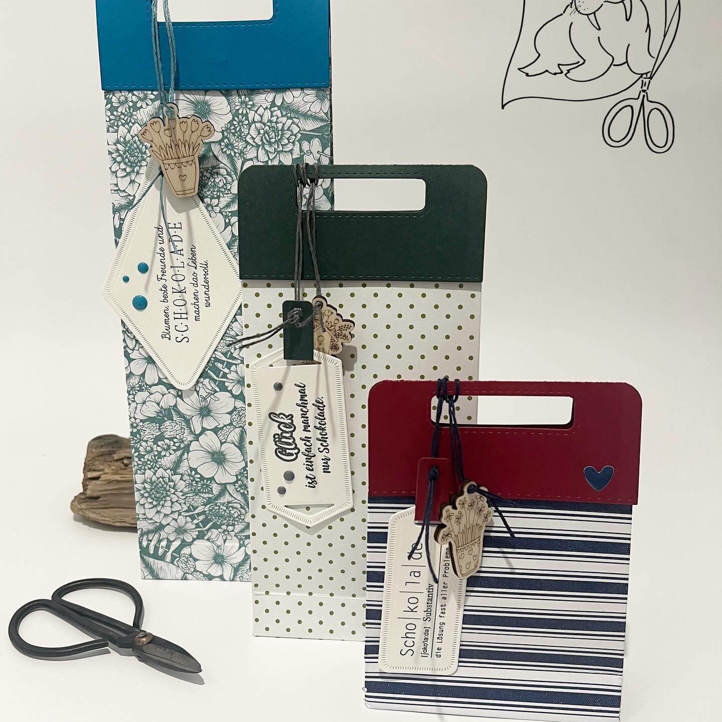 Drei gemusterte Geschenktüten mit Anhängern, die mit der Stanzschablone Griffe von Stanzenshop.de erstellt wurden, stehen aufrecht neben einer Schere und einem Holzartikel - ein tolles Bastelergebnis für Scrapbooking-Fans. Rechts oben ist eine Siegelzeichnung mit einer Schere zu sehen.