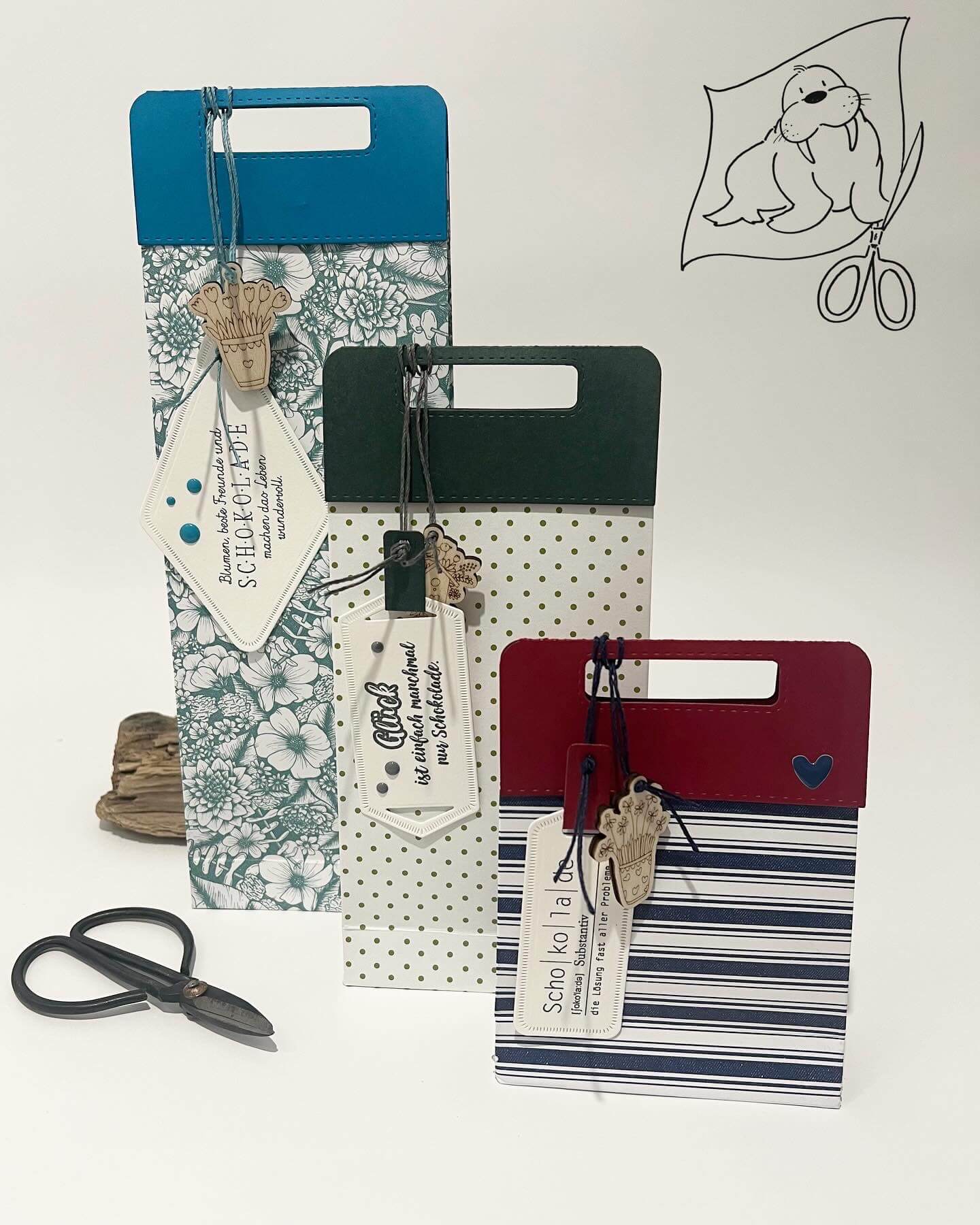 Drei gemusterte Geschenktüten mit Anhängern, die mit der Stanzschablone Griffe von Stanzenshop.de erstellt wurden, stehen aufrecht neben einer Schere und einem Holzartikel - ein tolles Bastelergebnis für Scrapbooking-Fans. Rechts oben ist eine Siegelzeichnung mit einer Schere zu sehen.