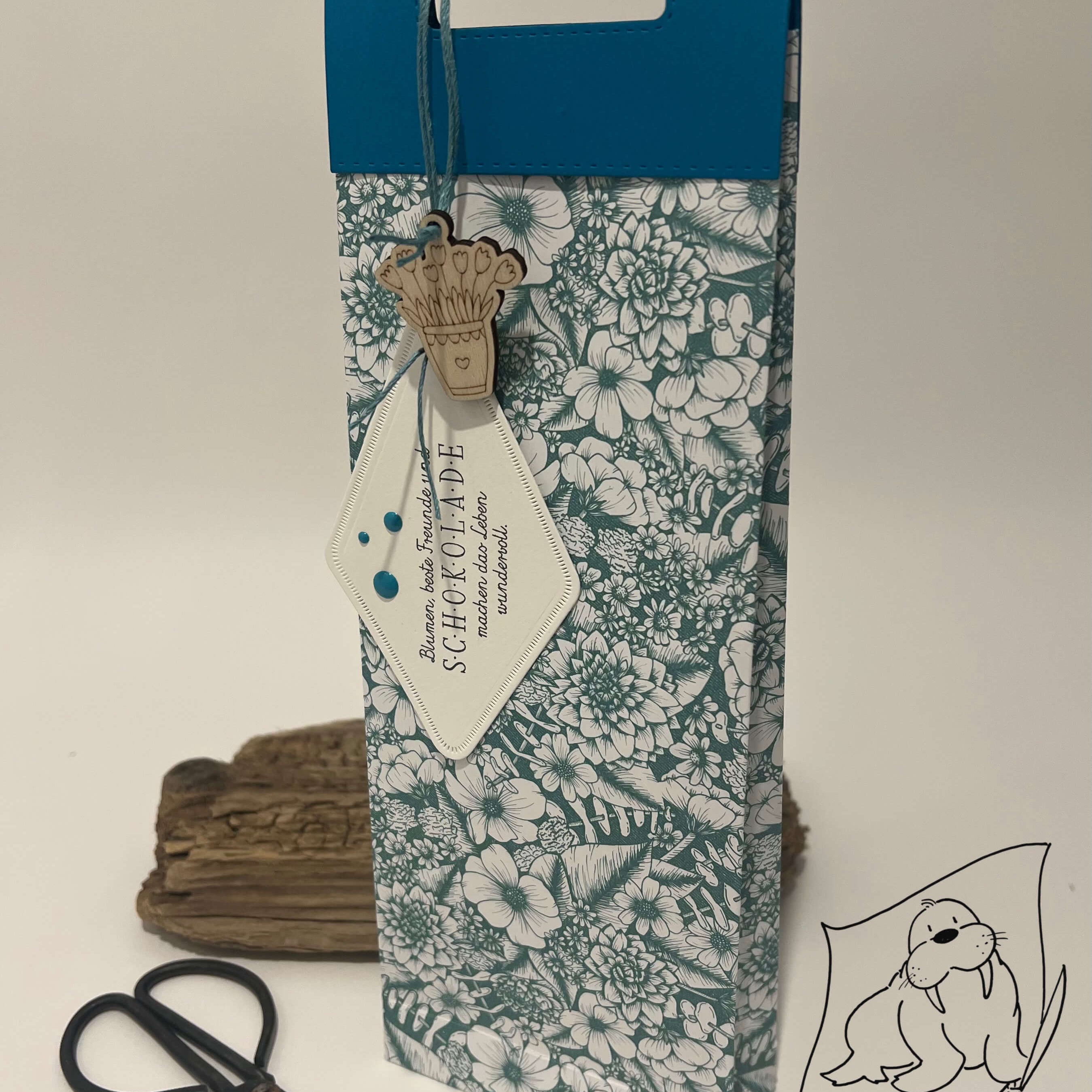 Große Geschenktasche mit Stanzschablone Griffe von Stanzenshop.de mit floralem Muster, blauem Deckel, Holzanhänger und weißem Etikett, aufrecht neben Schere und Treibholz auf neutralem Hintergrund.