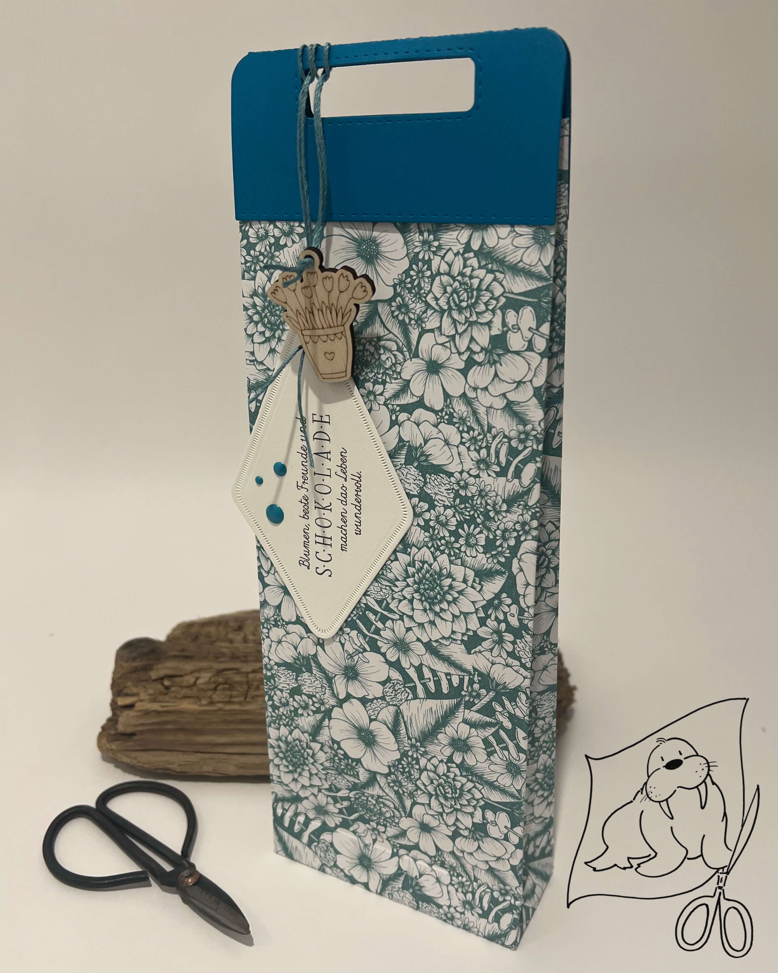 Große Geschenktasche mit Stanzschablone Griffe von Stanzenshop.de mit floralem Muster, blauem Deckel, Holzanhänger und weißem Etikett, aufrecht neben Schere und Treibholz auf neutralem Hintergrund.