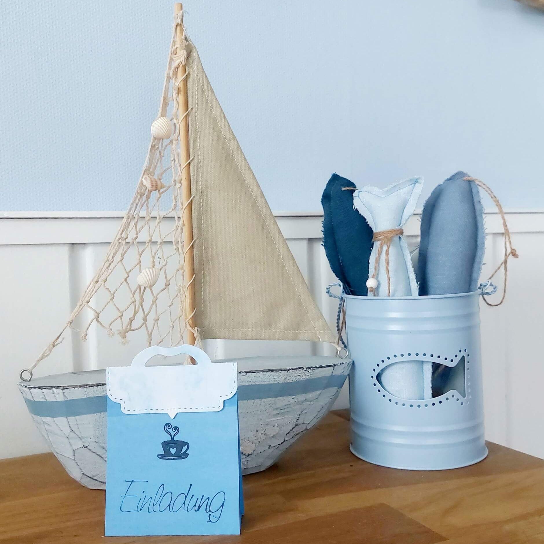 Ein Modellsegelboot, eine blaue Metallbox mit Stofffischen und eine blaue Einladungskarte aus der Stanzschablone Verschluss für Tüten von Stanzenshop.de sind auf einer Holzfläche vor einer hellblauen Wand angeordnet.