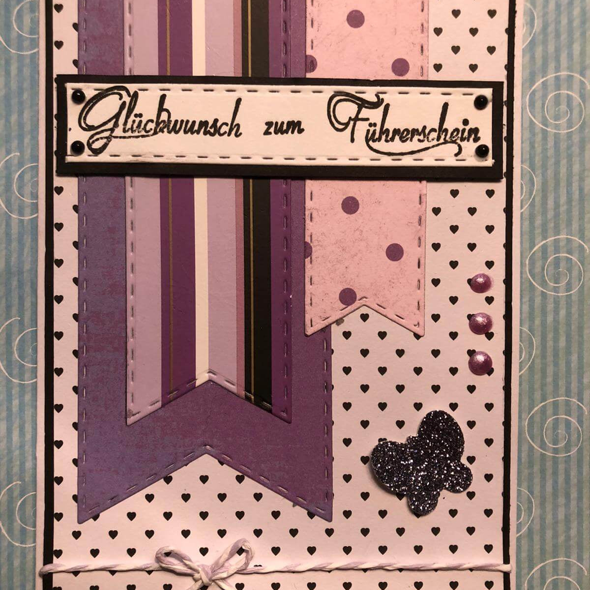 Handgefertigte Glückwunschkarte in Lila-Tönen mit "Glückwunsch zum Führerschein" und Bannern aus der Stanzschablone Fünf lange Wimpel von Stanzenshop.de, dazu ein glitzernder Schmetterling, Perlen, eine weiße Schleife und ein gemusterter Hintergrund.