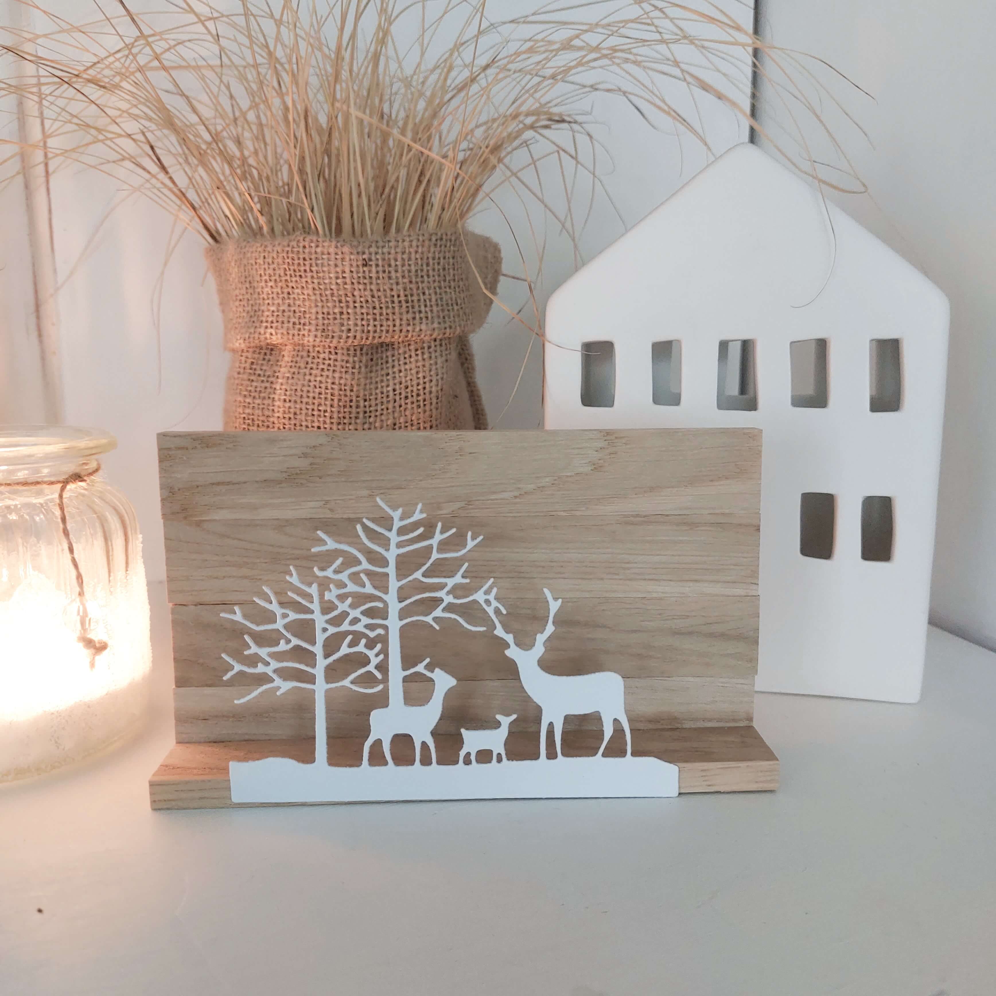 Die Stanzschablone "Winterlandschaft mit drei Rehen" von Stanzenshop.de ist als Holzständer mit weißen Rehen und Bäumen dargestellt, ideal für Basteln Deko, neben einer brennenden Kerze, Hausschmuck und getrocknetem Gras in einer Vase.