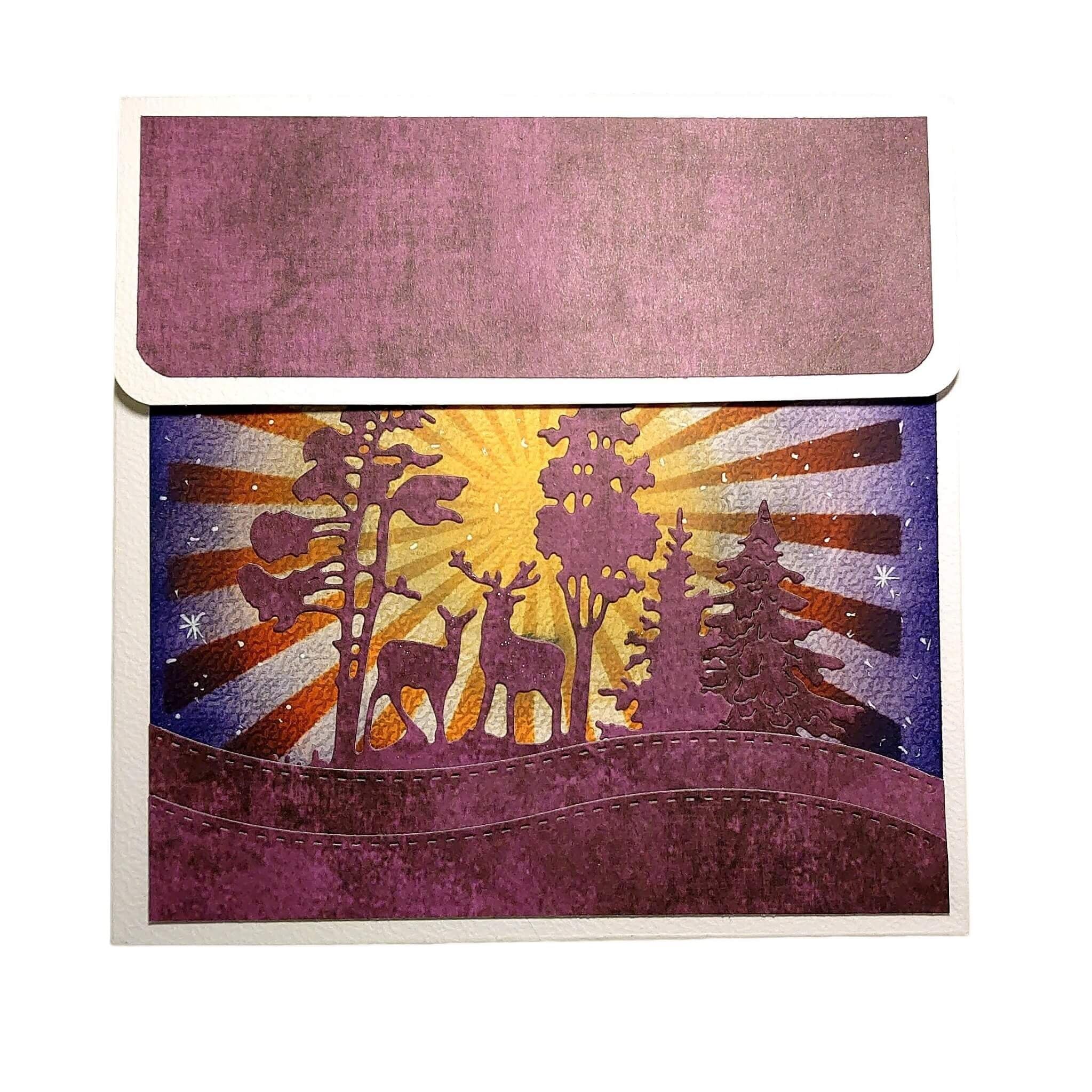 Eine Grußkarte mit einer lila-gelben Waldszene mit Rehen, Bäumen und einem Sonnenaufgang - perfekt für Basteln Karten oder in Kombination mit der Stanzschablone: Winterlandschaft im Halbkreis von Stanzenshop.de.