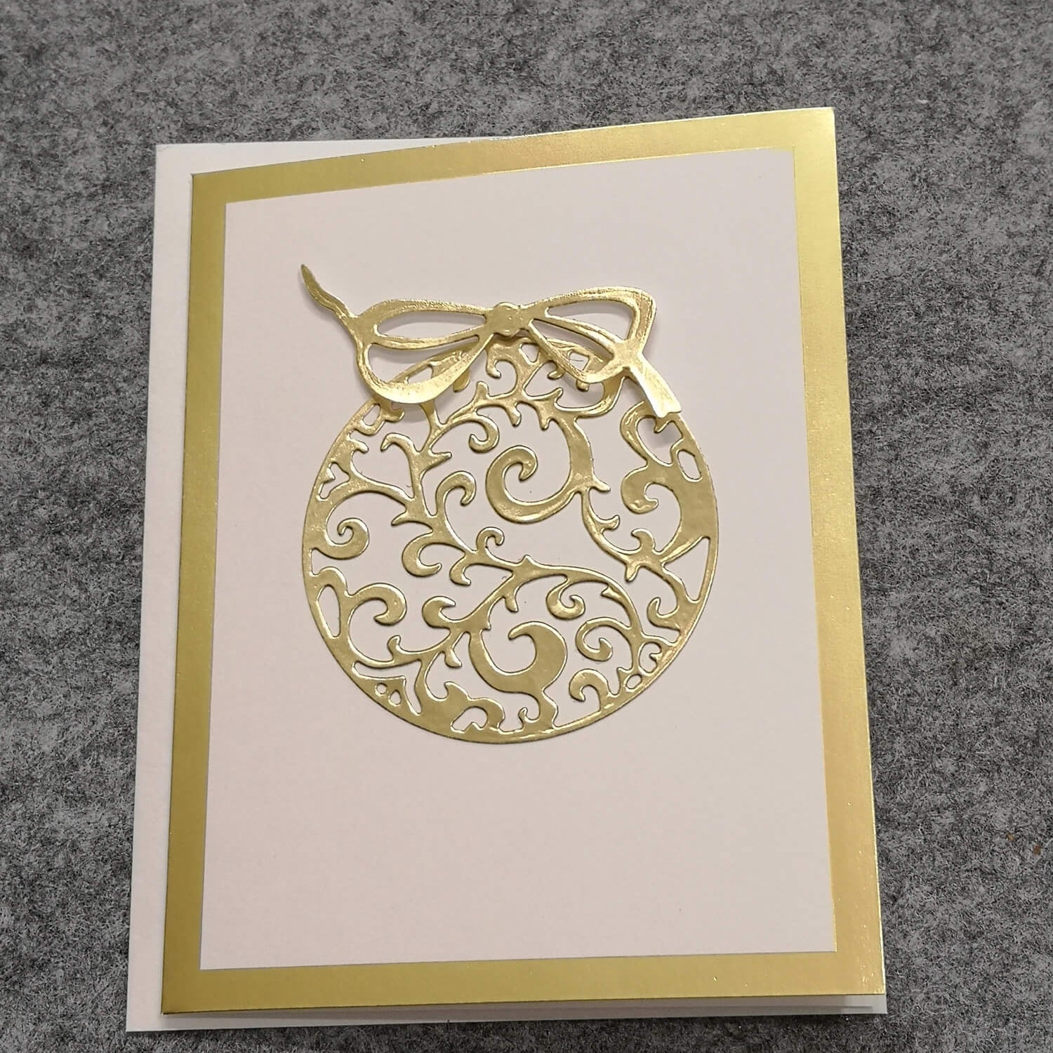 Eine weiße Grußkarte mit goldenem Rand zeigt ein festliches Ornamentmotiv, das von der "Stanzschablone" von Stanzenshop.de inspiriert ist: Zwei Weihnachtskugeln", auf grauem, strukturiertem Hintergrund - perfekt für Basteln Weihnachten.