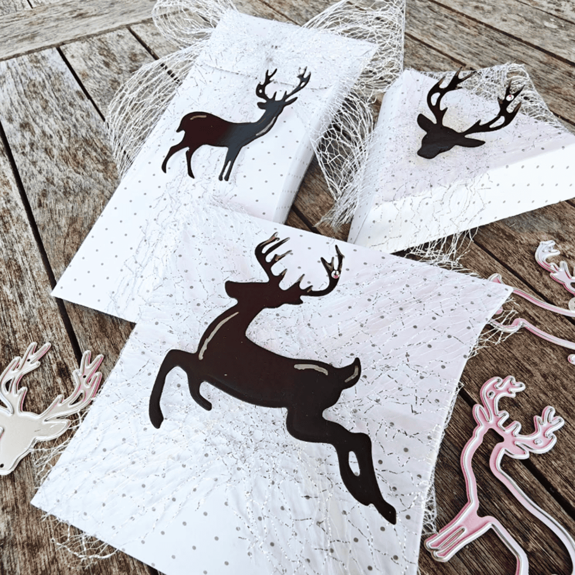 Drei weiße Geschenkboxen mit schwarzen Rentier-Silhouetten und Netz-Akzenten sitzen auf einer Holzfläche, umgeben von metallischen und rosafarbenen "Stanzschablone Drei Rentiere" von Stanzenshop.de-ideal für Weihnachts-Basteln.