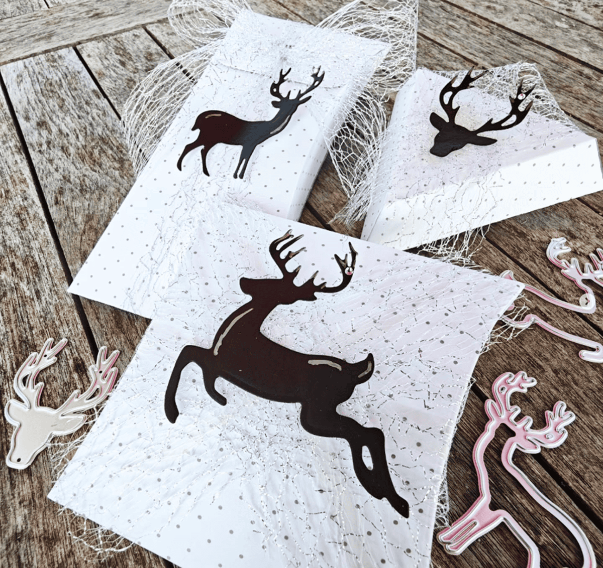 Drei weiße Geschenkboxen mit schwarzen Rentier-Silhouetten und Netz-Akzenten sitzen auf einer Holzfläche, umgeben von metallischen und rosafarbenen "Stanzschablone Drei Rentiere" von Stanzenshop.de-ideal für Weihnachts-Basteln.