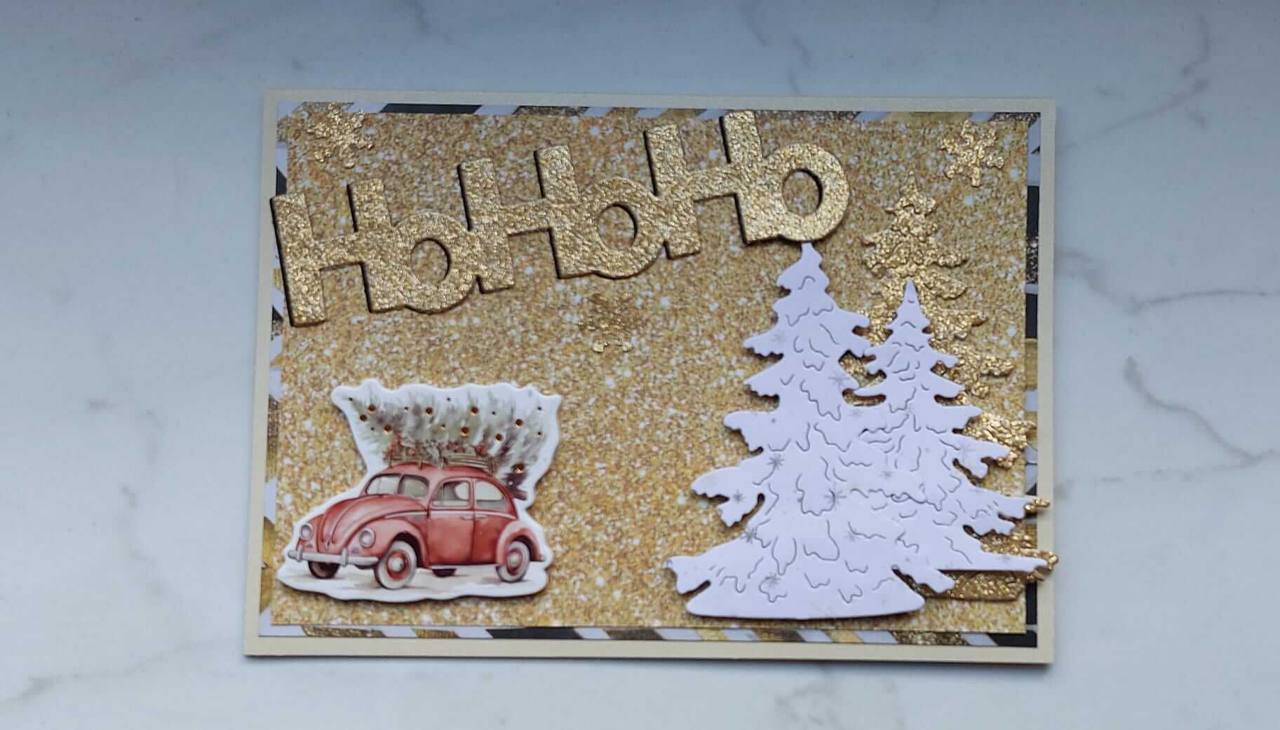 Gestalten Sie festliche Projekte mit dieser handgefertigten Weihnachtskarte mit dem Stanzschablone Schiftzug "hohoho" von Stanzenshop.de, einem glitzernden goldenen Hintergrund, einem roten Auto mit Baum und zwei weißen Tannen - ideal zum Basteln oder Scrapbooking.