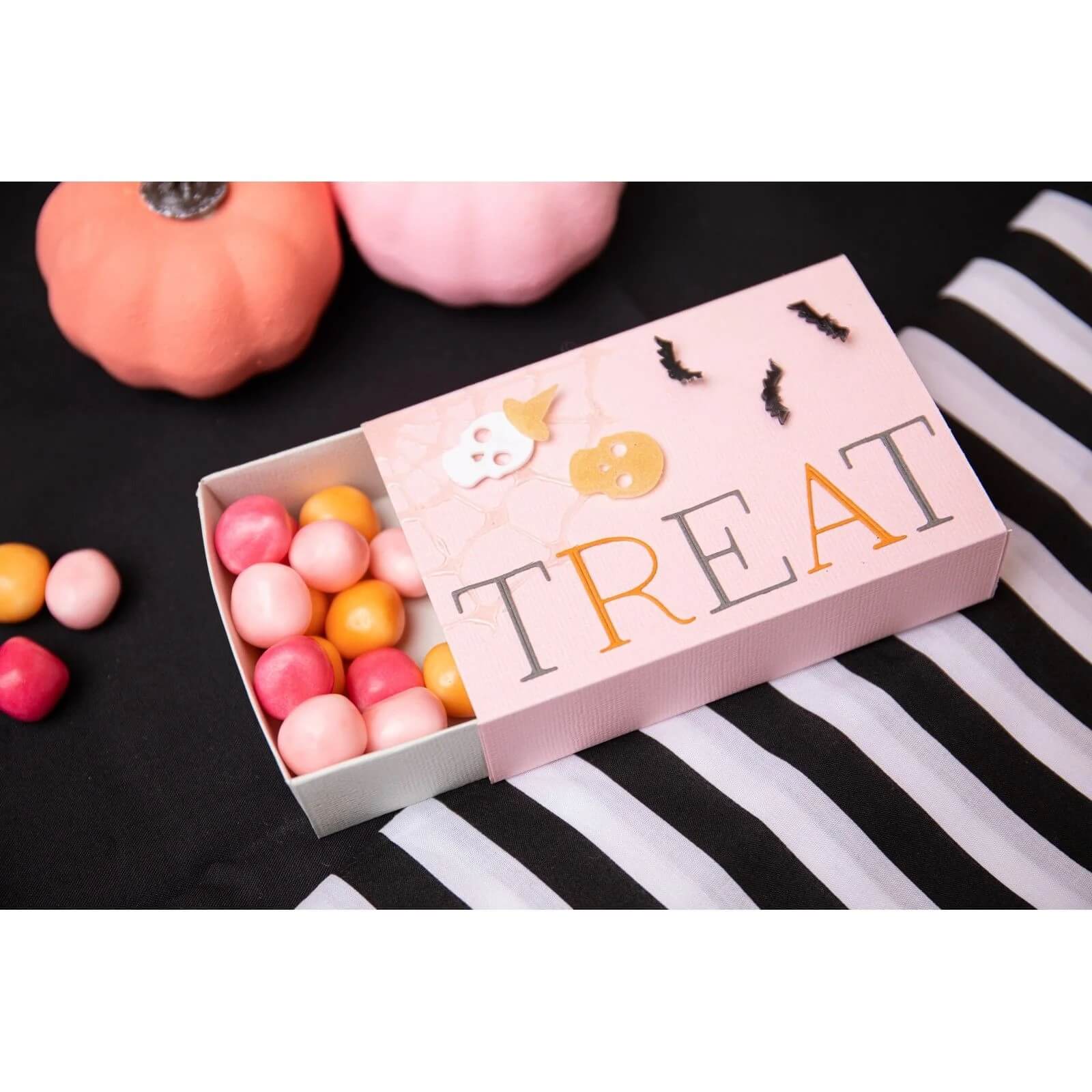 Eine rosafarbene "TREAT"-Schachtel, die mit den Sizzix 666453 - Thinlits Spooky Icons von Sizzix dekoriert wurde, sitzt teilweise geöffnet auf einem gestreiften Tuch und zeigt bunte Süßigkeiten im Inneren, mit zwei dekorativen Kürbissen in der Nähe.