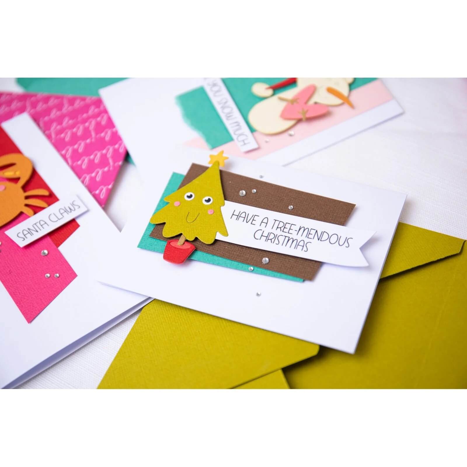 Eine handgefertigte Weihnachtskarte mit einem Papierbaum, die mit den Sizzix 666313 Thinlits mit Stamps-Christmas Characters hergestellt wurde, zeigt die Botschaft "Have a tree-mendous Christmas" auf einem Umschlag, der von festlichen Karten umgeben ist.