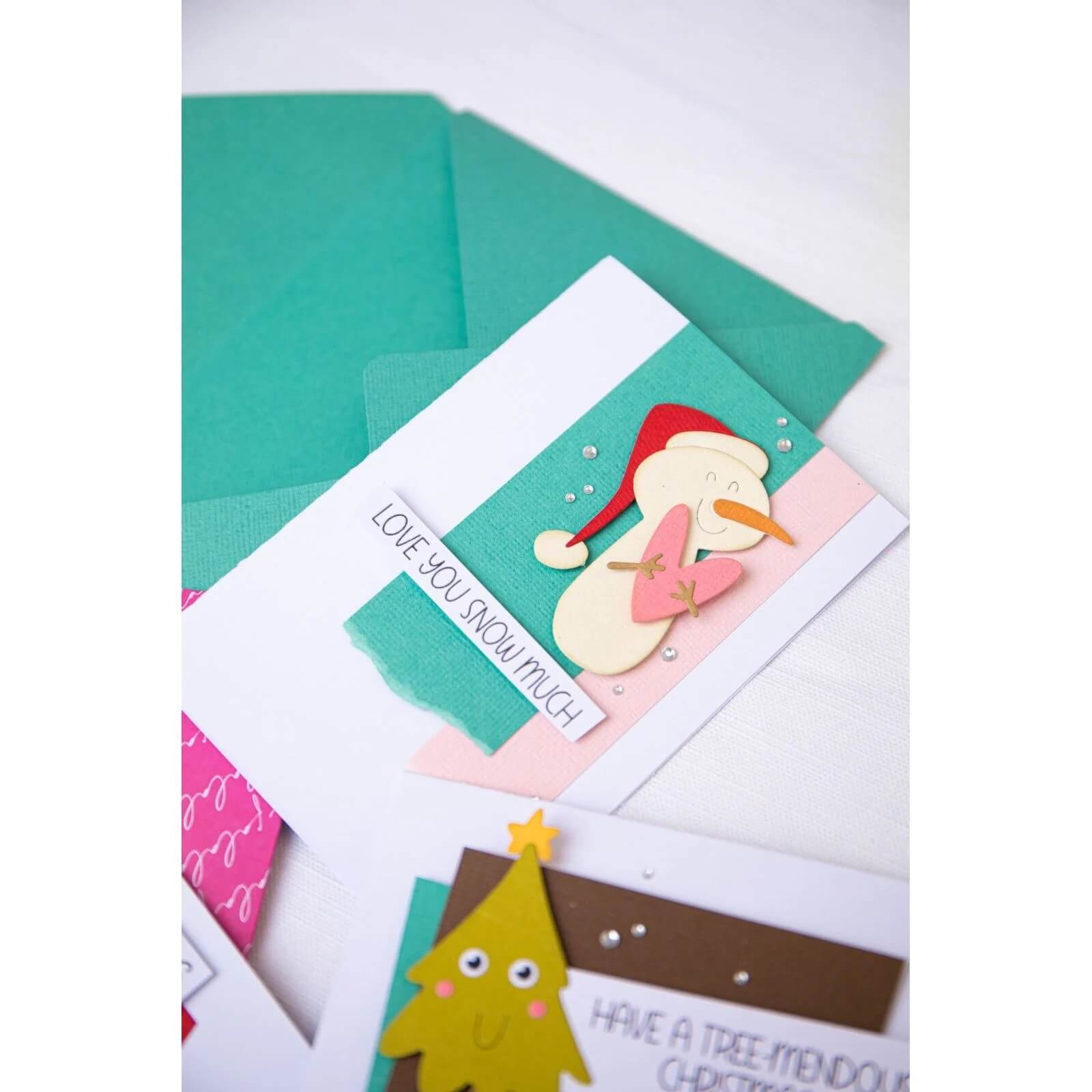 Eine handgefertigte Weihnachtskarte, die mit den Sizzix 666313 Thinlits with Stamps - Christmas Characters (von Sizzix) hergestellt wurde, zeigt einen Schneemann mit roter Weihnachtsmannmütze und der Aufschrift "LOVE YOU SNOW MUCH", der auf einem grünen Umschlag inmitten von festlichem Bastelmaterial ruht.
