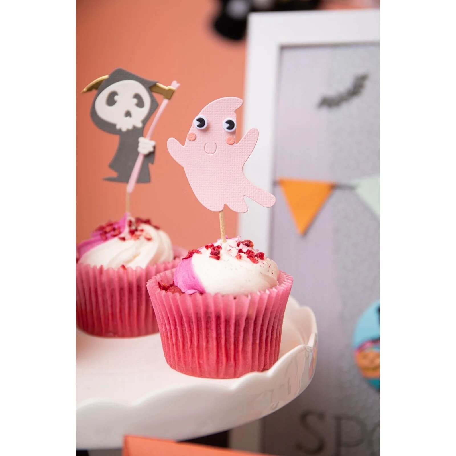 Zwei Cupcakes mit rosafarbenem Zuckerguss sind mit Papieraufsätzen - einem Cartoon-Gespenst und einem Sensenmann - versehen, die mit den Sizzix 666315 - Thinlits mit Stamps Spooky Friends von Sizzix hergestellt wurden. Festliche Banner vervollständigen diese Papierbastelprojekte zum Thema Halloween.