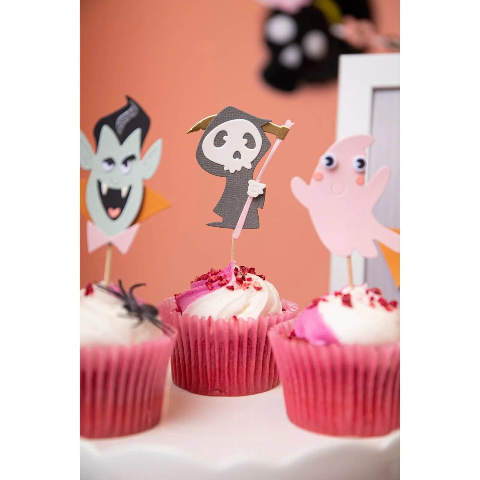 Drei Cupcakes mit rosafarbenem Deckblatt und weißem Zuckerguss werden mit Halloween-Figuren verziert, die mit den Sizzix 666315 Thinlits mit den Stempeln Spooky Friends von Sizzix hergestellt wurden: ein Vampir, ein Sensenmann und ein Geist. Ein Cupcake ist mit einer Plastikspinne verziert.