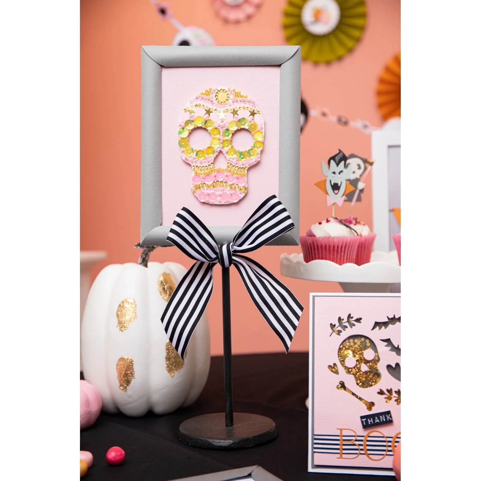 Ein gerahmter Pailletten-Totenkopf auf einem Ständer mit einer schwarz-weiß gestreiften Schleife steht inmitten von Halloween-Dekor - weißer Kürbis, Muffins und Akzente, die mit den Sizzix 666453 - Thinlits Spooky Icons von Sizzix hergestellt wurden.