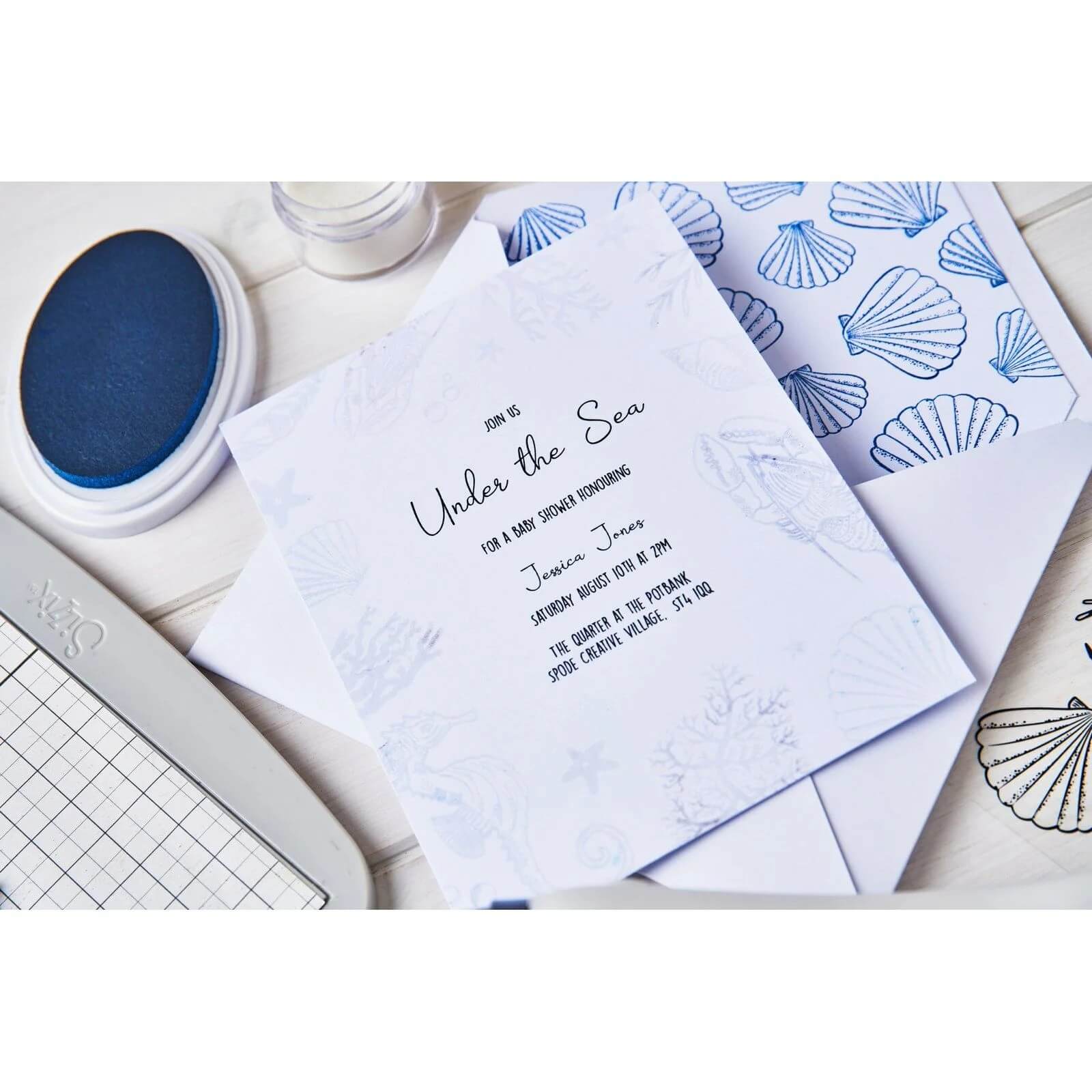 Eine weiße Einladungskarte "Under the Sea" mit Meeresmotiven, die mit dem Sizzix 666669 Clear Stamp Set Ocean Elements (15 Stück) von Sizzix hergestellt wurde, liegt auf einem Tisch neben einem blauen Umschlag mit Muschelmuster, einem Stempelkissen und einer Schneidematte.