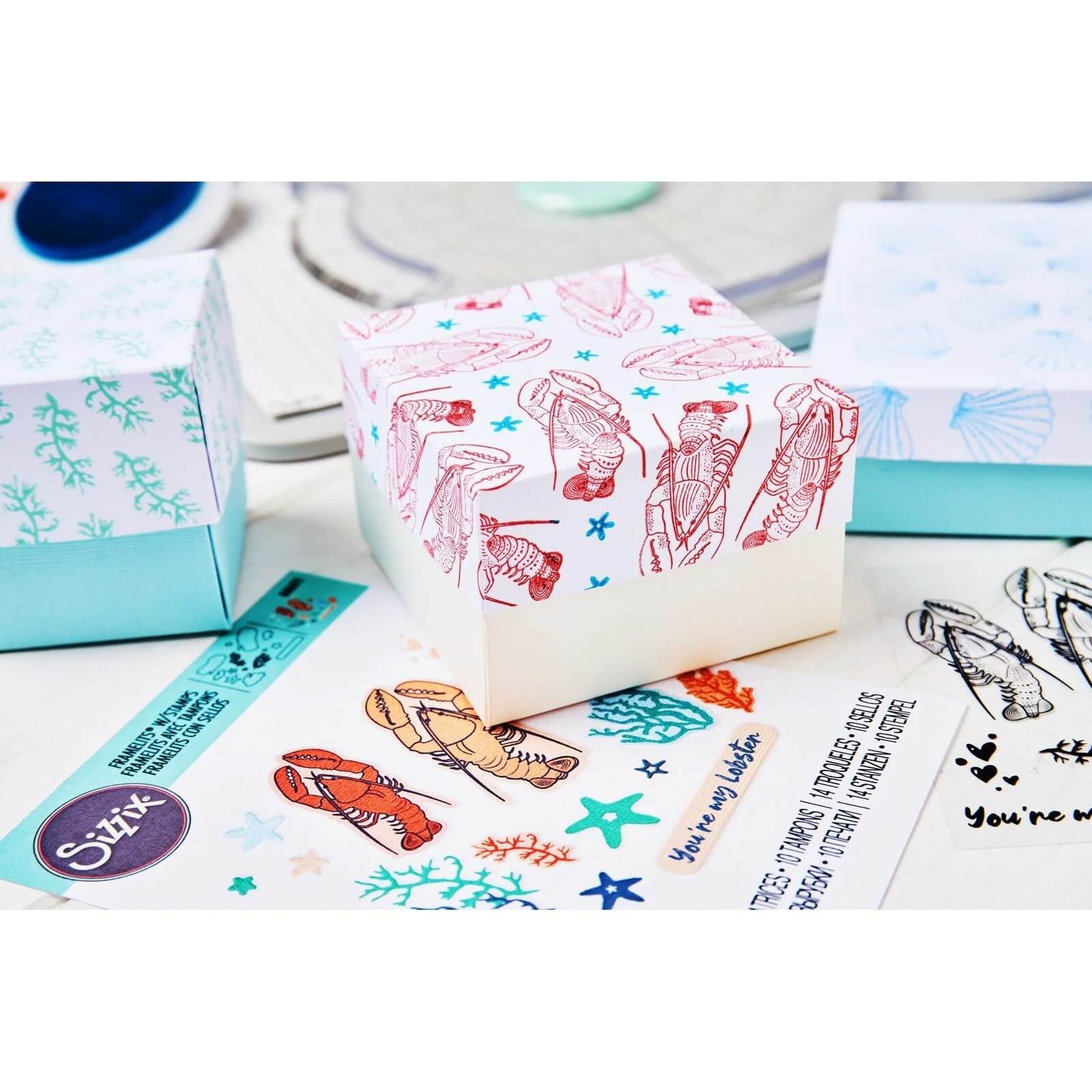 Eine Geschenkschachtel mit Muschel- und Seepferdchenmotiven, die mit dem Sizzix 666669 Clear Stamp Set Ocean Elements (15 Stück) gestempelt wurden, befindet sich zwischen pastellfarbenen Schachteln und Aufklebern mit Meeresmotiven auf einer weißen Fläche.