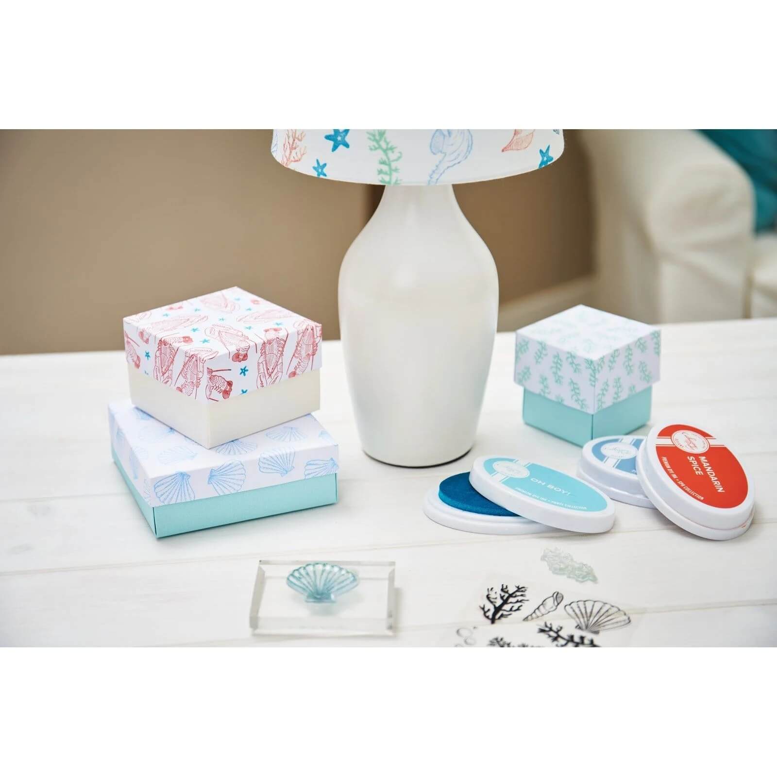 Zwei gemusterte Geschenkboxen, eine weiße Lampe mit dekorativem Schirm und das Framelits Set 666671 Framelits mit Stempeln "You're my Lobster" (24 Stück) von Sizzix werden auf einem weißen Tisch präsentiert - ideal, um herzliche Momente zu gestalten.