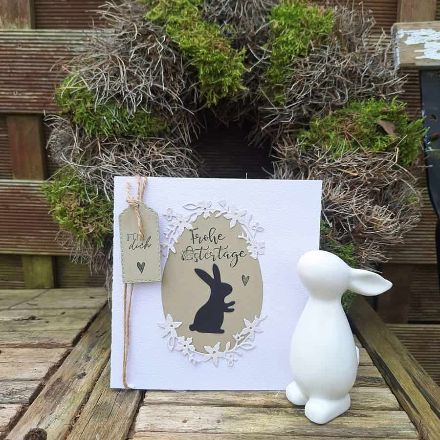 Eine weiße Osterkarte mit schwarzem Hasen und deutschem Text steht neben einem weißen Keramikhasen vor der Stanzschablone Blumenkranz von Stanzenshop.de, ausgestellt auf Holz - die ideale Inspiration für Basteln-Fans.
