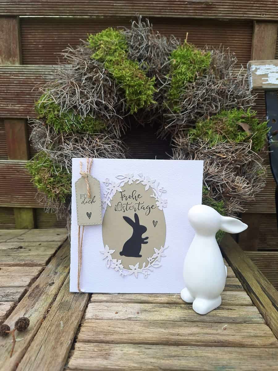 Eine weiße Osterkarte mit schwarzem Hasen und deutschem Text steht neben einem weißen Keramikhasen vor der Stanzschablone Blumenkranz von Stanzenshop.de, ausgestellt auf Holz - die ideale Inspiration für Basteln-Fans.