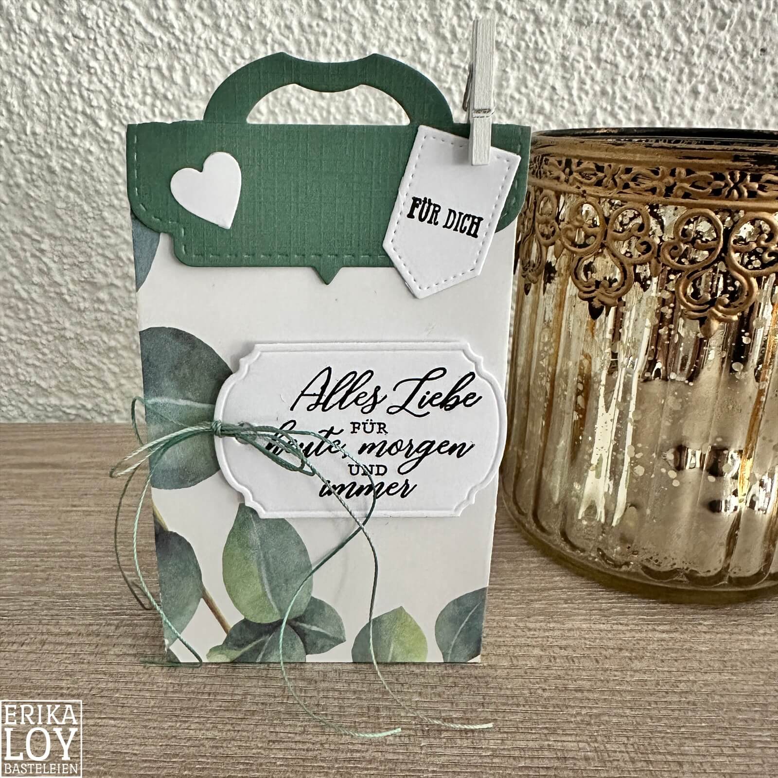 Die "Stanzschablone Verschluss für Tüten" von Stanzenshop.de wird als Geschenkbox mit grünem Henkel, Herzausschnitt und "Für dich"-Anhänger mit Blattmotiv - perfekt zum Basteln - neben einem Goldglas auf Holz gezeigt.
