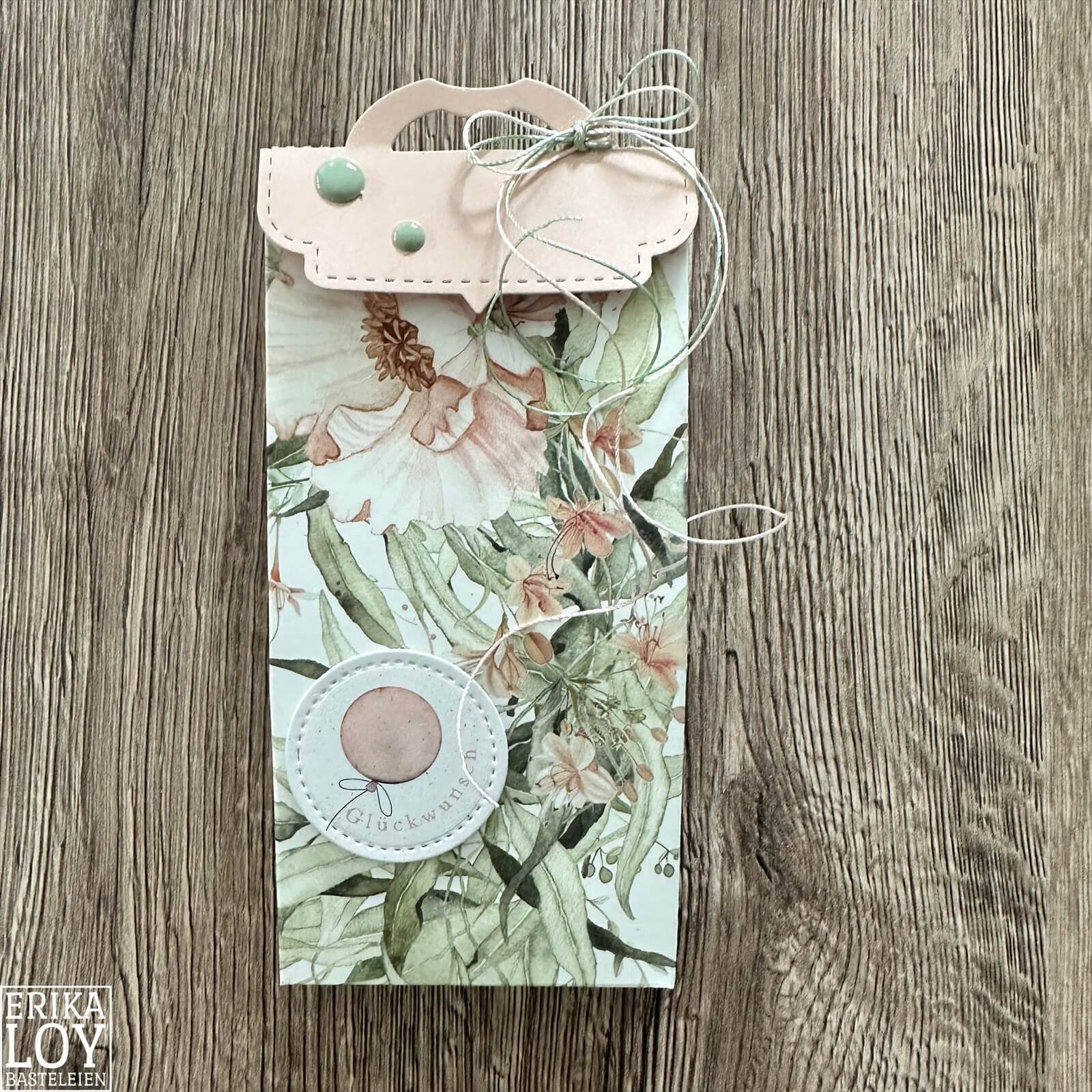 Eine Geschenktüte mit floralen Mustern, pastellfarbenen Details und einem runden Anhänger mit der Aufschrift "Glückwunsch" auf Holz - die perfekte Inspiration für Basteln mit der Stanzschablone Verschluss für Tüten von Stanzenshop.de.