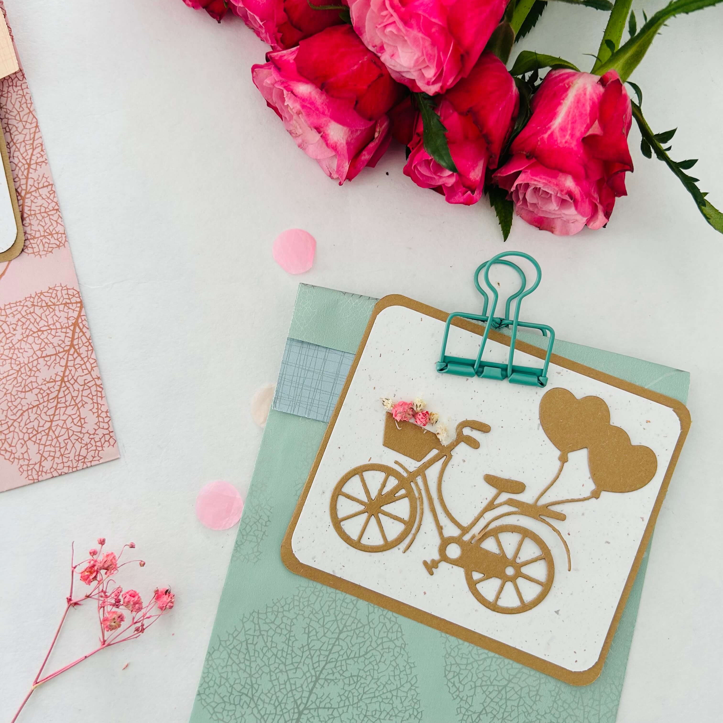 Die Stanzenshop.de-Grußkarte "Stanzschablone Fahrrad mit Luftballons", ideal zum Basteln oder Scrapbooking, wird in einen pastellblauen Umschlag gesteckt und neben rosa Rosen auf eine weiße Fläche gelegt.