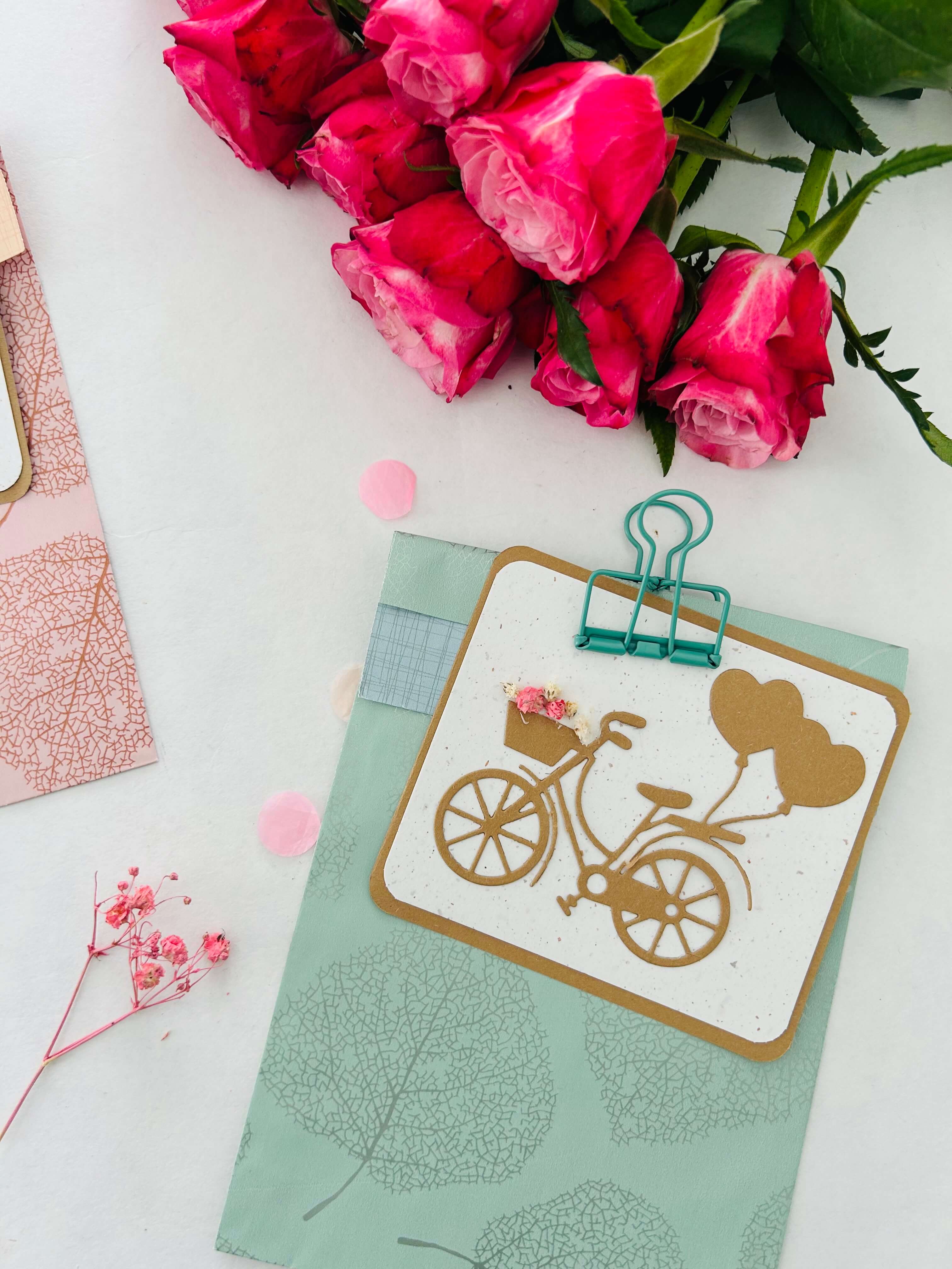 Die Stanzenshop.de-Grußkarte "Stanzschablone Fahrrad mit Luftballons", ideal zum Basteln oder Scrapbooking, wird in einen pastellblauen Umschlag gesteckt und neben rosa Rosen auf eine weiße Fläche gelegt.
