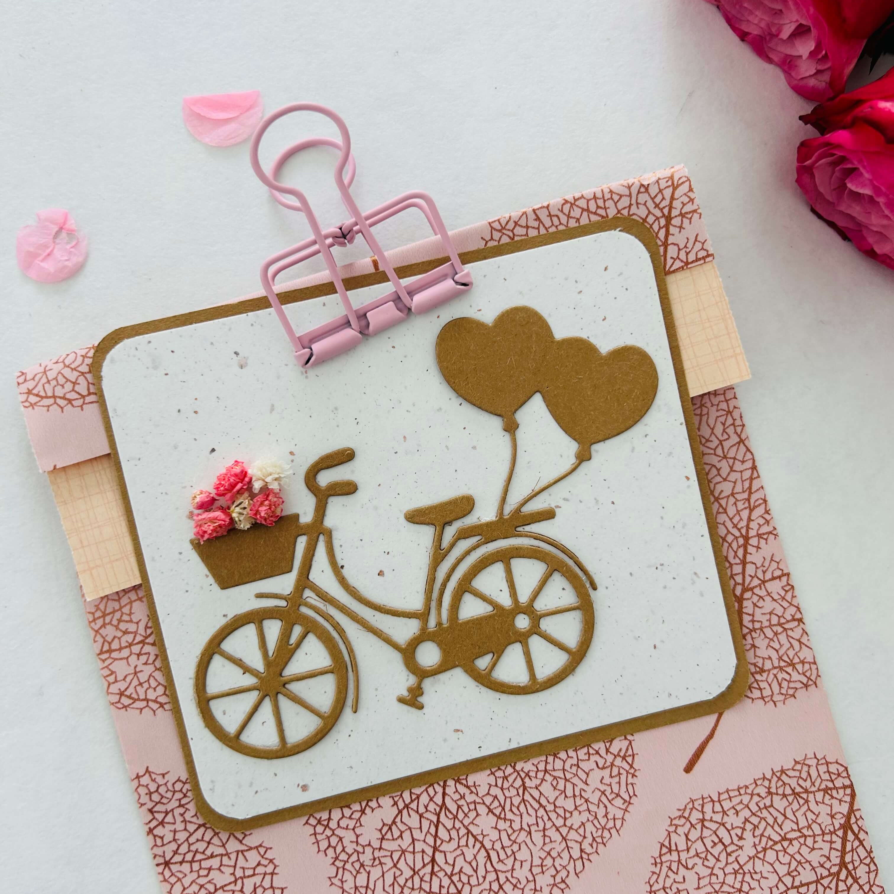 Die Stanzschablone Fahrrad mit Luftballons" von Stanzenshop.de zeigt ein braunes Fahrrad mit herzförmigen Luftballons und einem Blumenkorb, ideal für Scrapbooking oder Basteln, aufgeklebt auf einen rosa gemusterten Umschlag mit rosa Rosen.