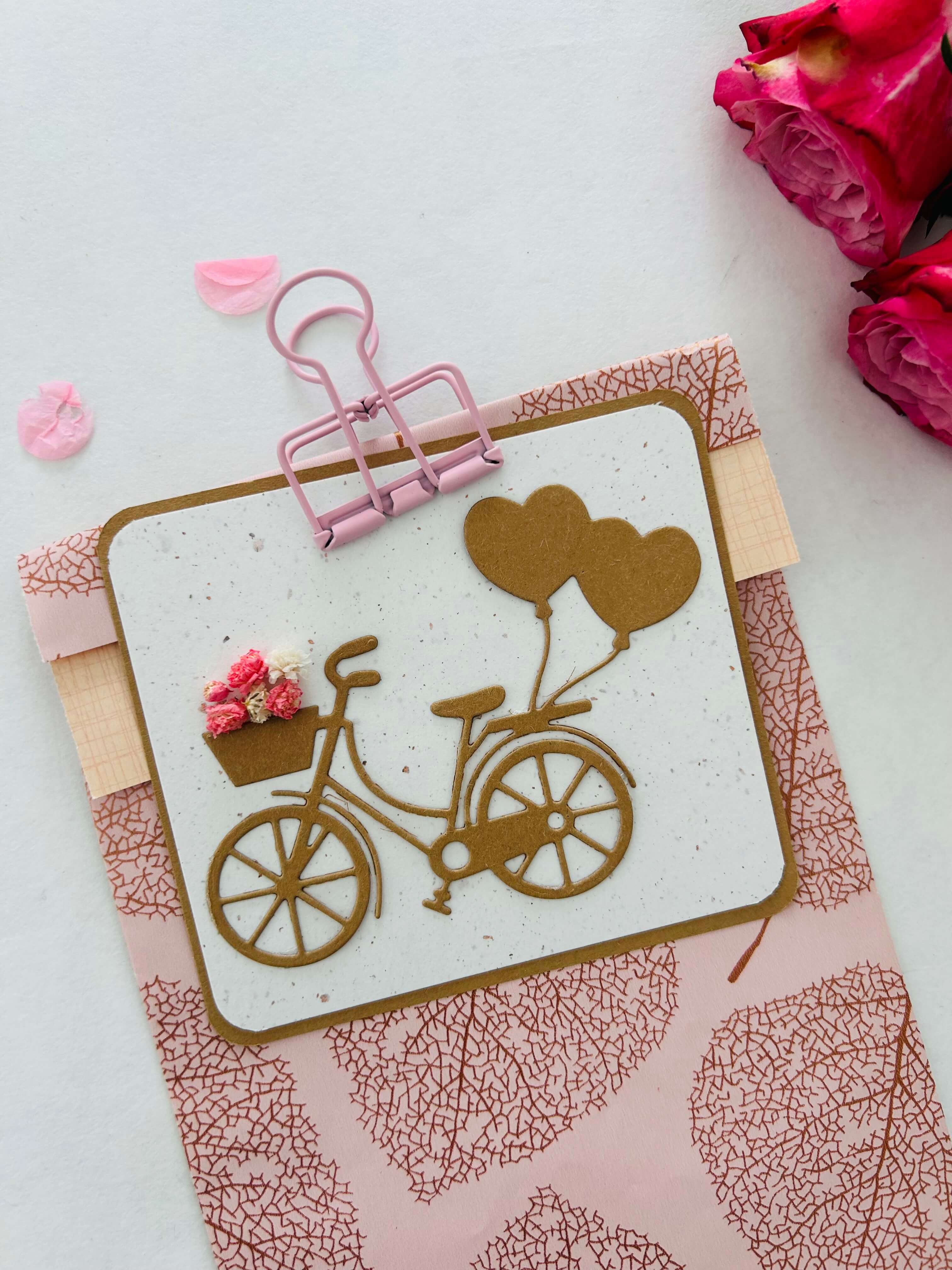 Die Stanzschablone Fahrrad mit Luftballons" von Stanzenshop.de zeigt ein braunes Fahrrad mit herzförmigen Luftballons und einem Blumenkorb, ideal für Scrapbooking oder Basteln, aufgeklebt auf einen rosa gemusterten Umschlag mit rosa Rosen.