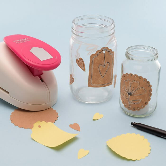 Zwei Gläser mit braunen Papieranhängern und Herzen sind umgeben von Florence Kraftklebepapier quadratisch 30,5x30,5cm (10 Stück), ausgeschnittenen Formen, einer anhängerförmigen Stanze und einem schwarzen Marker auf hellblauem Untergrund.