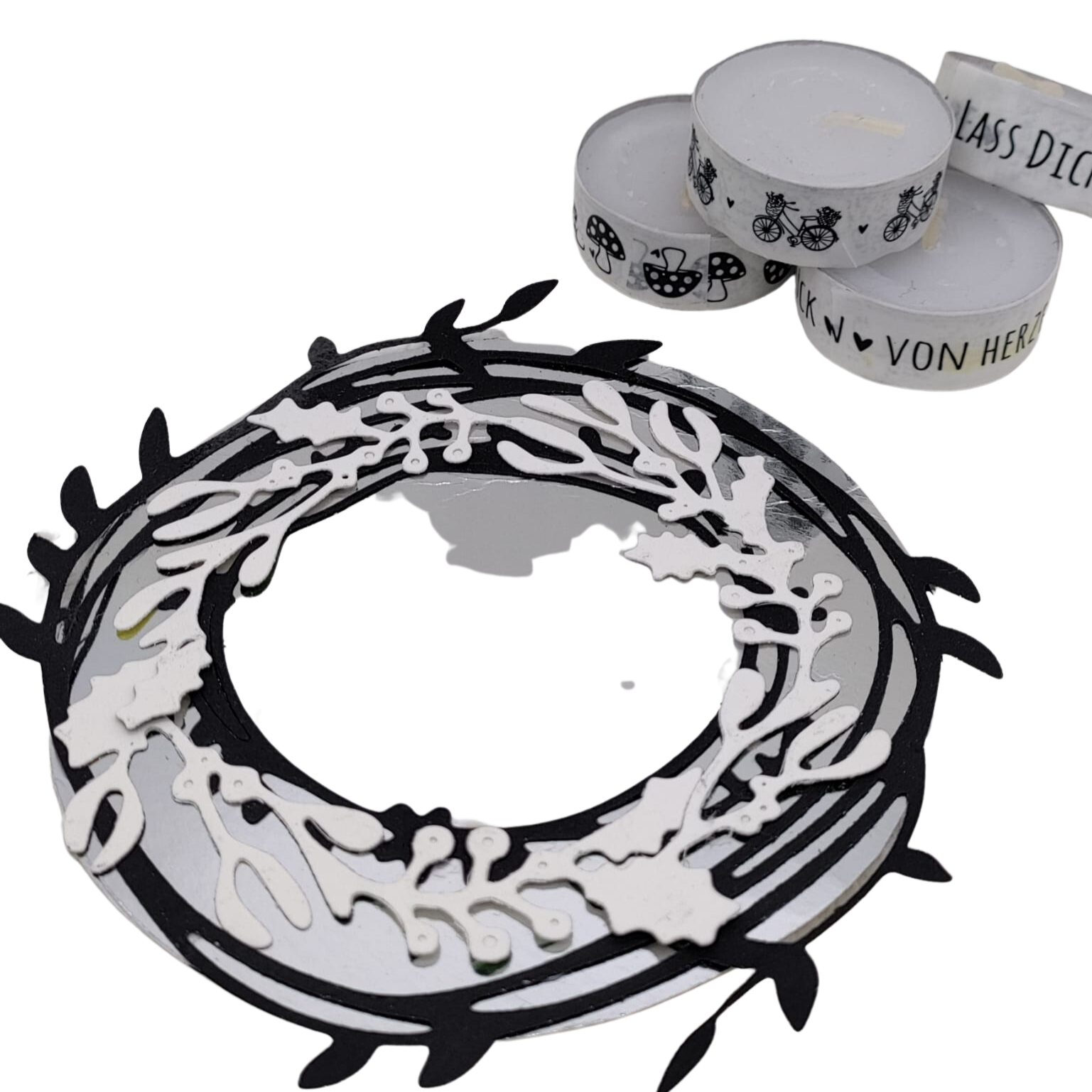 Die Stanzschablone: Kranz mit Beeren von Stanzenshop.de ist ein schwarz-weißer Papierkranz mit ausgeschnittenen Details, ideal zum Basteln oder Scrapbooking, abgebildet neben drei weißen Teelichtkerzen mit schwarzem Aufdruck.