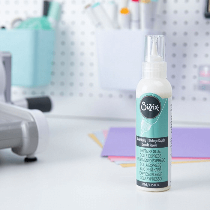 Eine Flasche Sizzix 664576 - Making Essential Express Kleber 120ml von Sizzix steht auf einem Schreibtisch neben farbigem Papier, mit Bastelwerkzeugen, die im Hintergrund verschwimmen - ideal für detaillierte Projekte, die Präzision erfordern.
