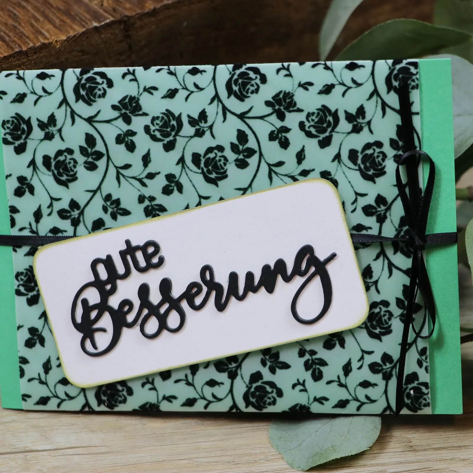 Eine 4 x 9 cm große grüne Karte von Mundart Stempel zeigt ein schwarzes Blumenmuster, ein weißes Etikett mit dem Schriftzug "Gute Besserung", das mit der Stanzschablone der Marke geschnitten wurde, und ist mit einem schwarzen Band gebunden.
