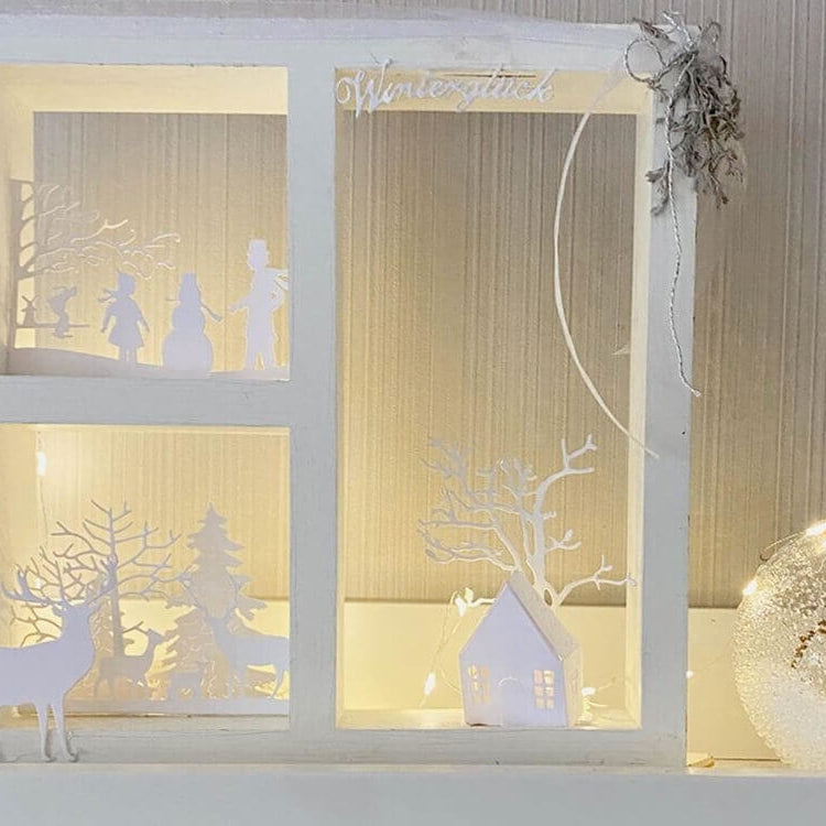 Eine Schattenbox aus weißem Holz, die mit dem Stanzschablone Bastelset Kleines Haus von Stanzenshop.de hergestellt wurde, zeigt winterliche Papierszenen - Bäume, Schneemann, Rentier - die von warmen Lichtern neben einem glitzernden Ornament beleuchtet werden.