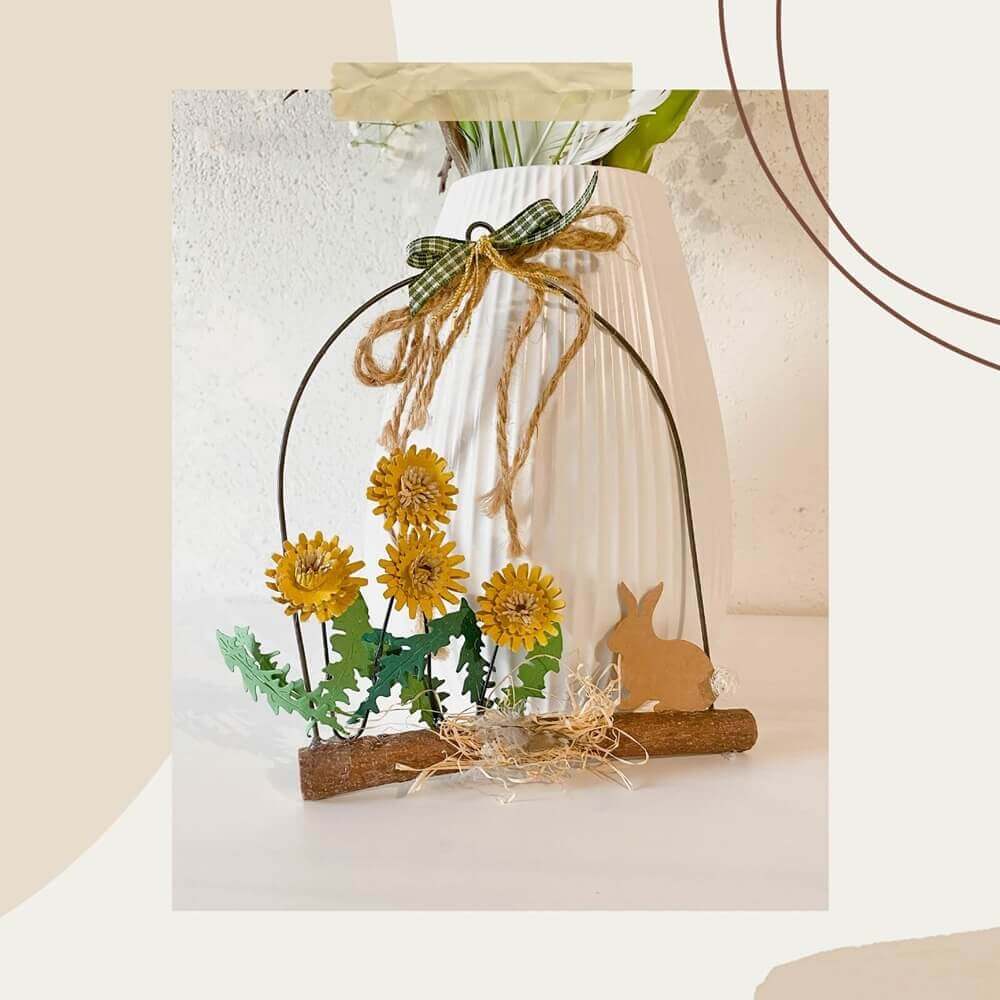 Das Stanzschablone Bastelset Löwenzahn von Stanzenshop.de zeigt gelbe Pusteblumen, Grünzeug und eine braune Kaninchensilhouette auf einem Baumstamm mit Schleife - ideal für Scrapbooking - vor einer weißen, strukturierten Vase.