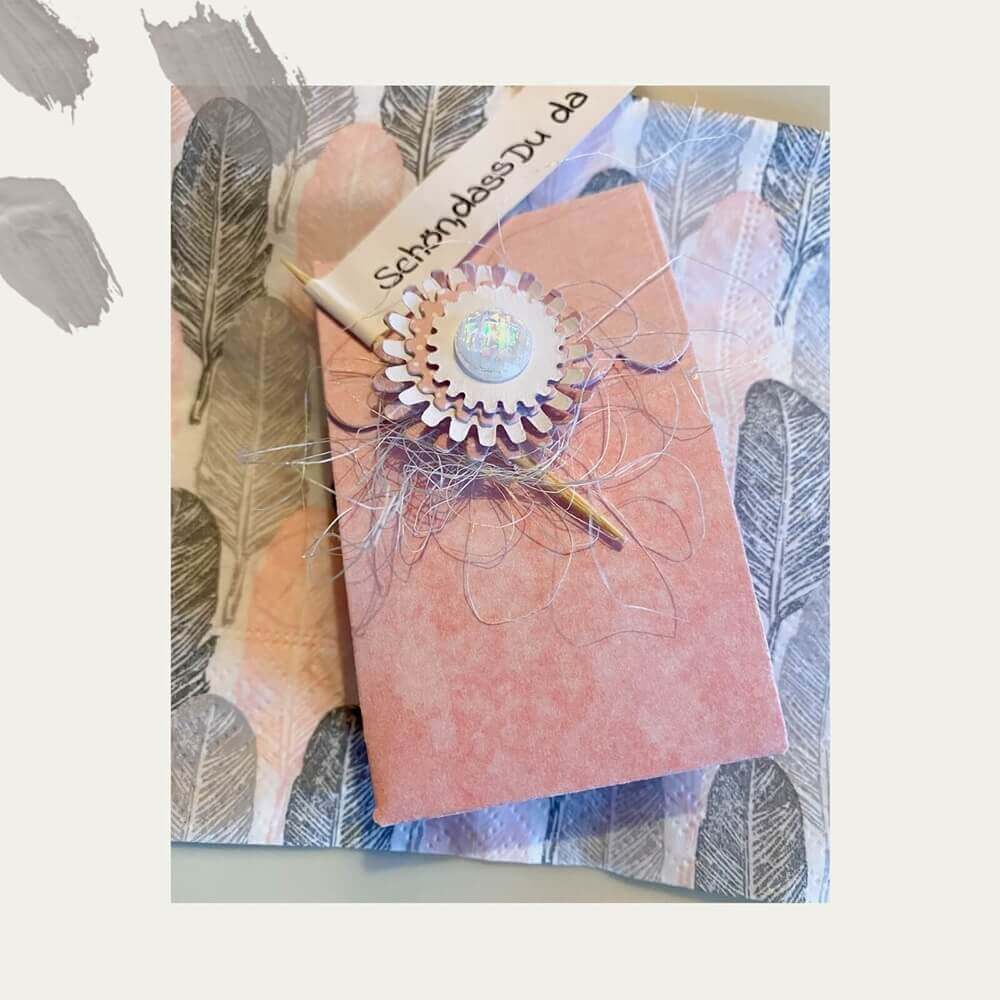 Eine rosafarbene Geschenkbox mit einem Blumenornament ruht auf einem federgemusterten Hintergrund neben einer deutschen Karte - ideal für Scrapbooking oder als Teil der Stanzschablone Bastelset Löwenzahn von Stanzenshop.de.
