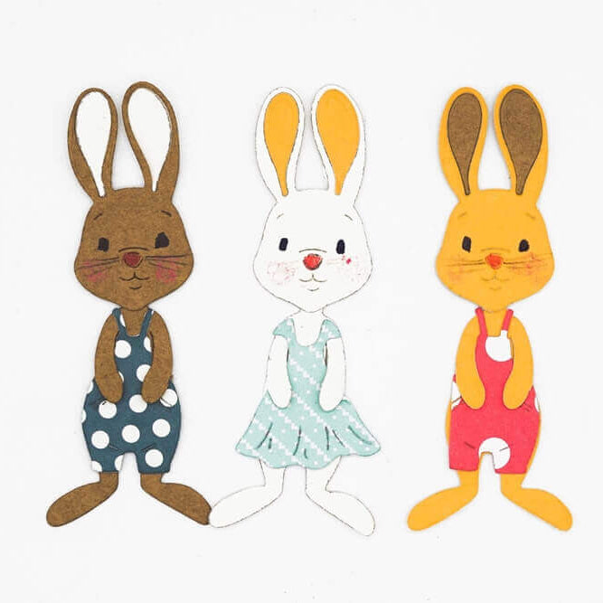 Die Stanzenshop.de Stanzschablone: Bastelset Osterhase zeigt drei Hasenausschnitte in bunten Kleidern, ideal für Scrapbooking oder Basteln. Braun mit marineblauen Punkten, weiß mit blauem Kleid und orange mit rotem Kleid auf weißem Hintergrund.