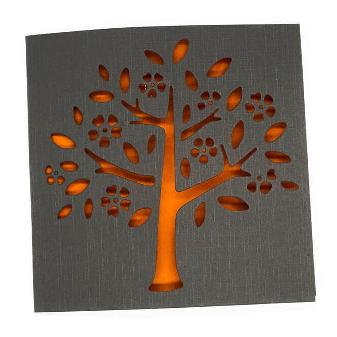 Eine quadratische schwarze Karte mit Baum-, Blüten- und Blattausschnitten leuchtet in orangefarbenem Licht - ideal für Scrapbooking oder Basteln mit der "Stanzschablone Baum mit Blüten und Blättern" von Stanzenshop.de.
