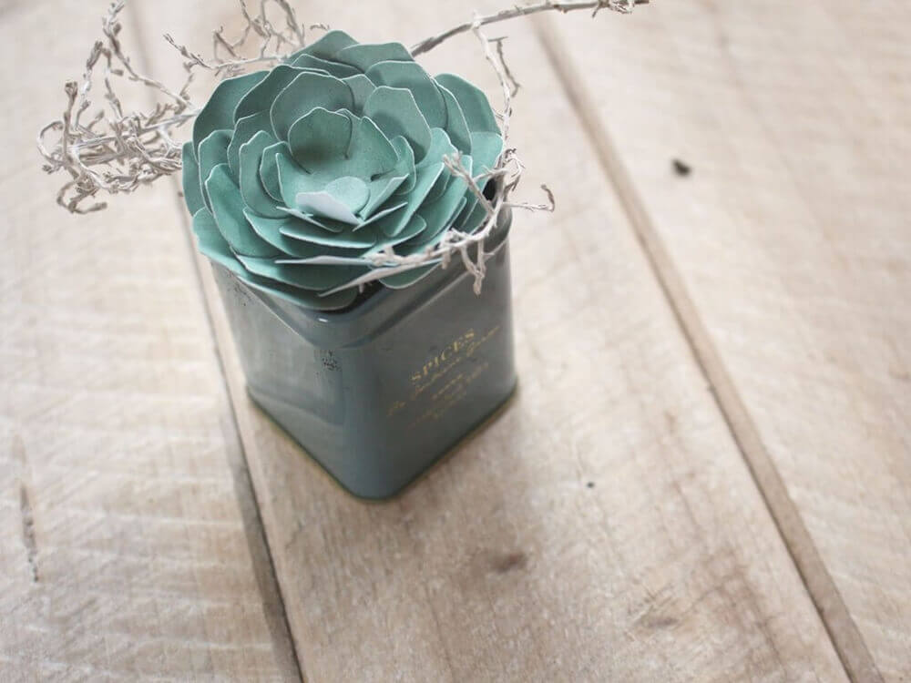 Eine grüne Papiersukkulente aus Stanzschablone: Blume aus mehreren Einzelteilen von Stanzenshop.de und weiße Zweige sind in einer quadratischen Dose arrangiert - perfekt für Scrapbooking oder Bastel Stanzen Projekte auf Holz.