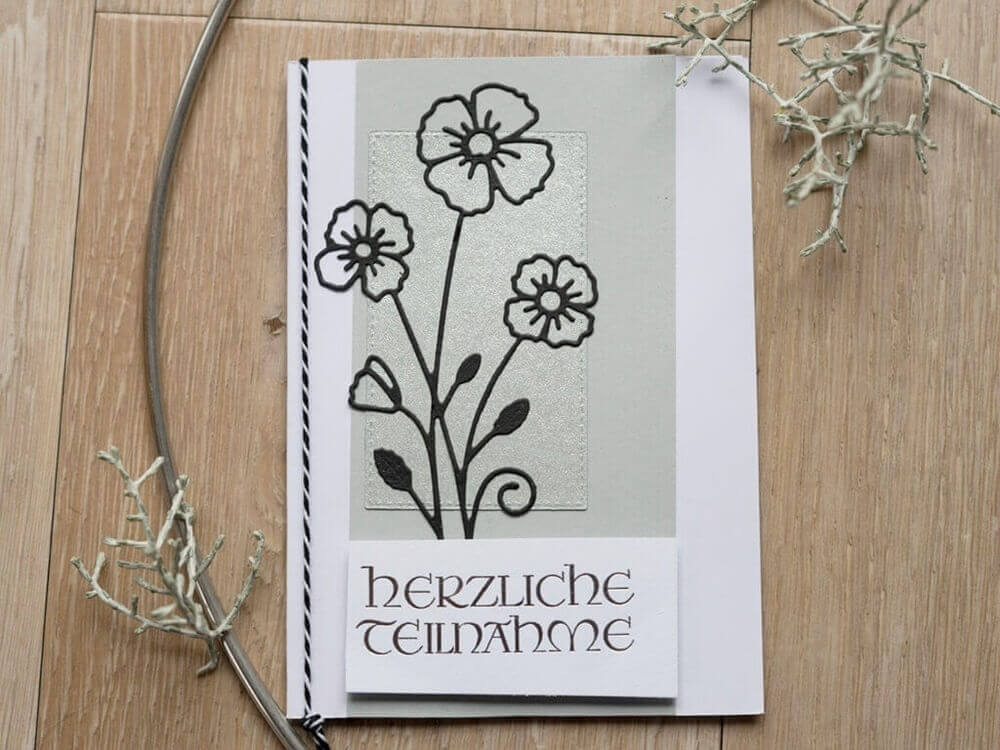 Eine Beileidskarte mit schwarzen Blumen, die mit der "Stanzschablone Blume mit drei Blüten" von Stanzenshop.de auf hellgrauem Hintergrund gestaltet wurde, mit der Aufschrift "Herzliche Anteilnahme" auf Deutsch. Die Karte ist auf Holz gelegt und mit dekorativen Zweigen umgeben.